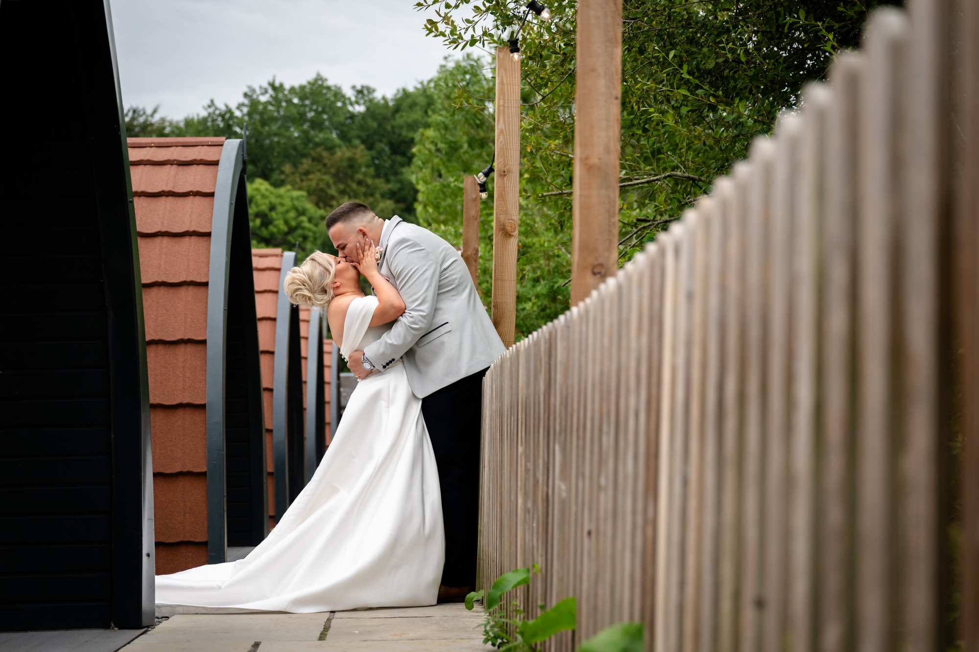 An intimate moment with the groom kissing the bride on the forehead, leaning against a wooden fence at Hanbury Barns.