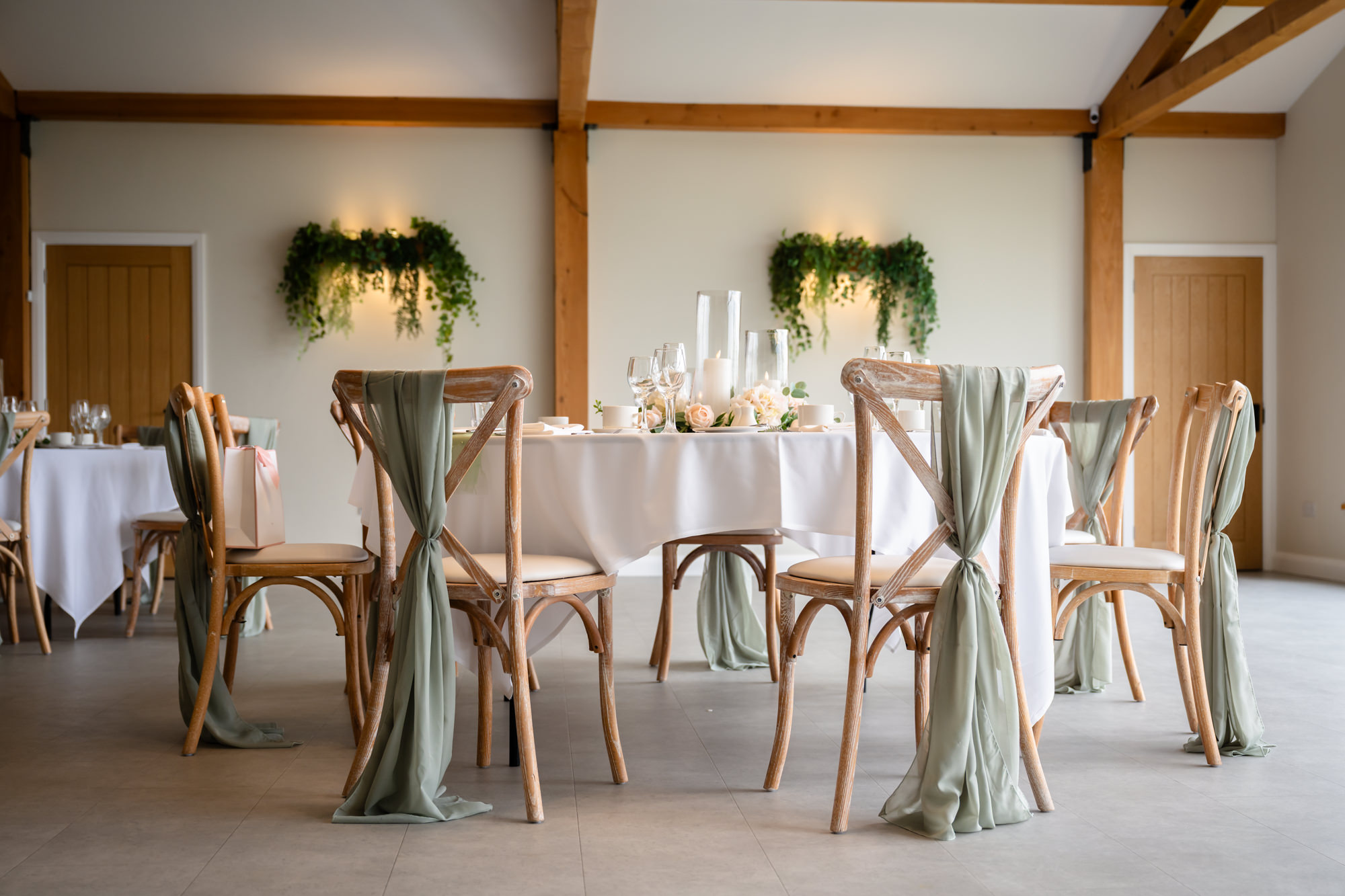 A Hanbury Barns wedding view of the reception room focused on the wooden cross-back chairs decorated with white ribbons awaiting guests.