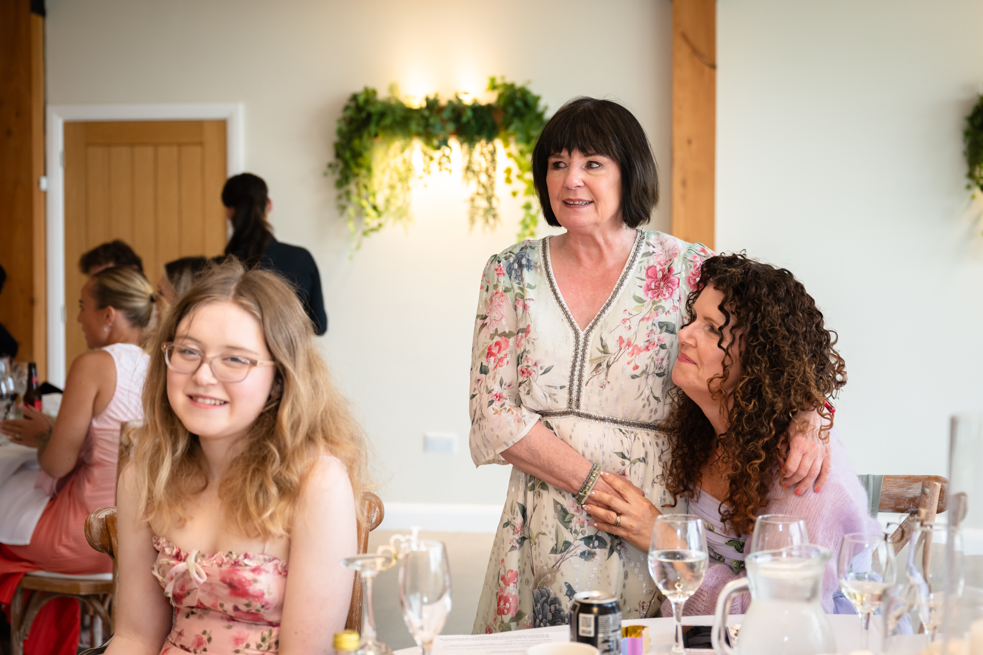 A woman embracing a guest with short dark hair, smiling while standing next to a young girl inside Hanbury Barns.
