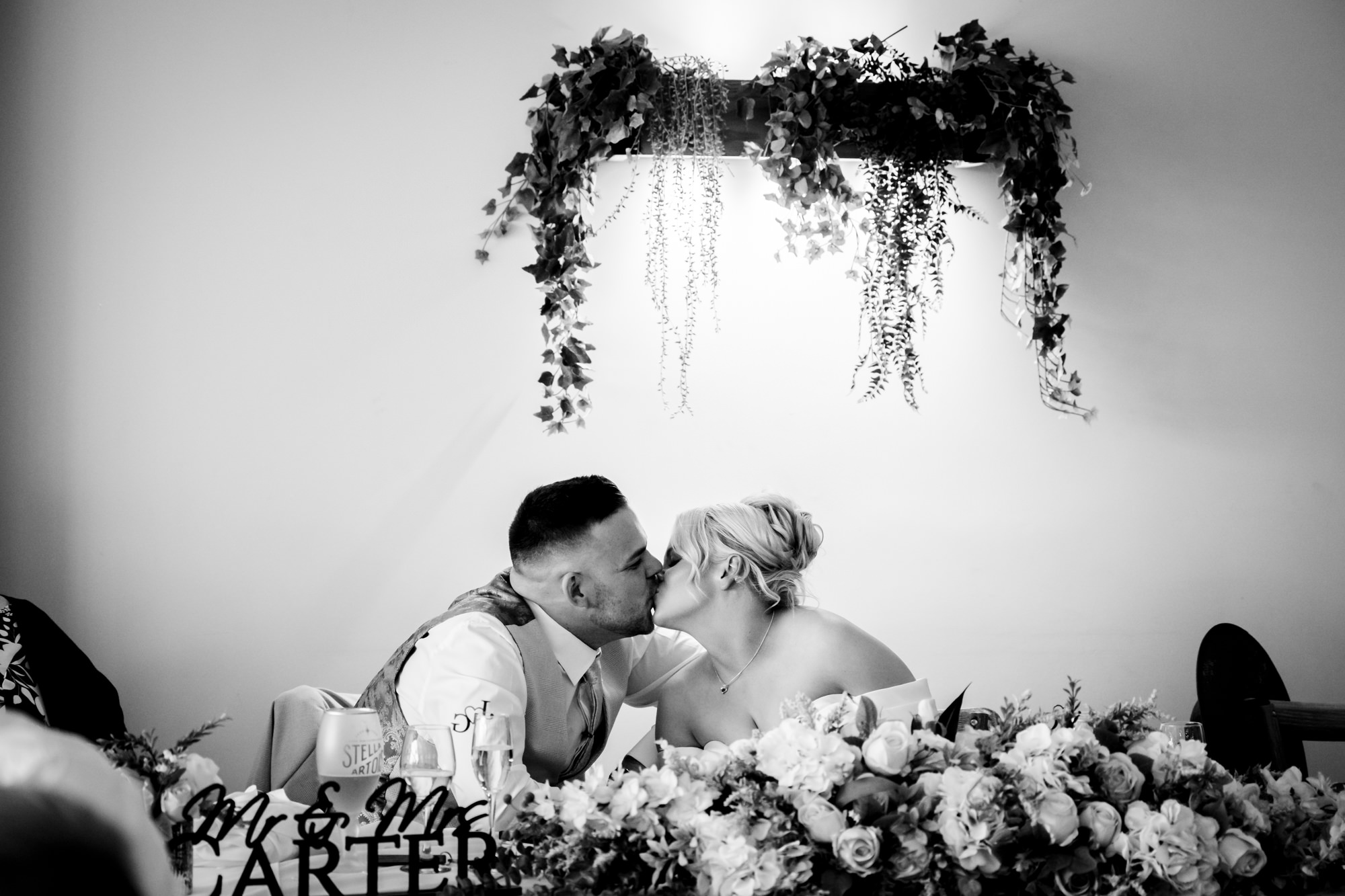 A black and white photo of the bride and groom kissing across the head table, decorated with a large floral arrangement at Hanbury Barns.