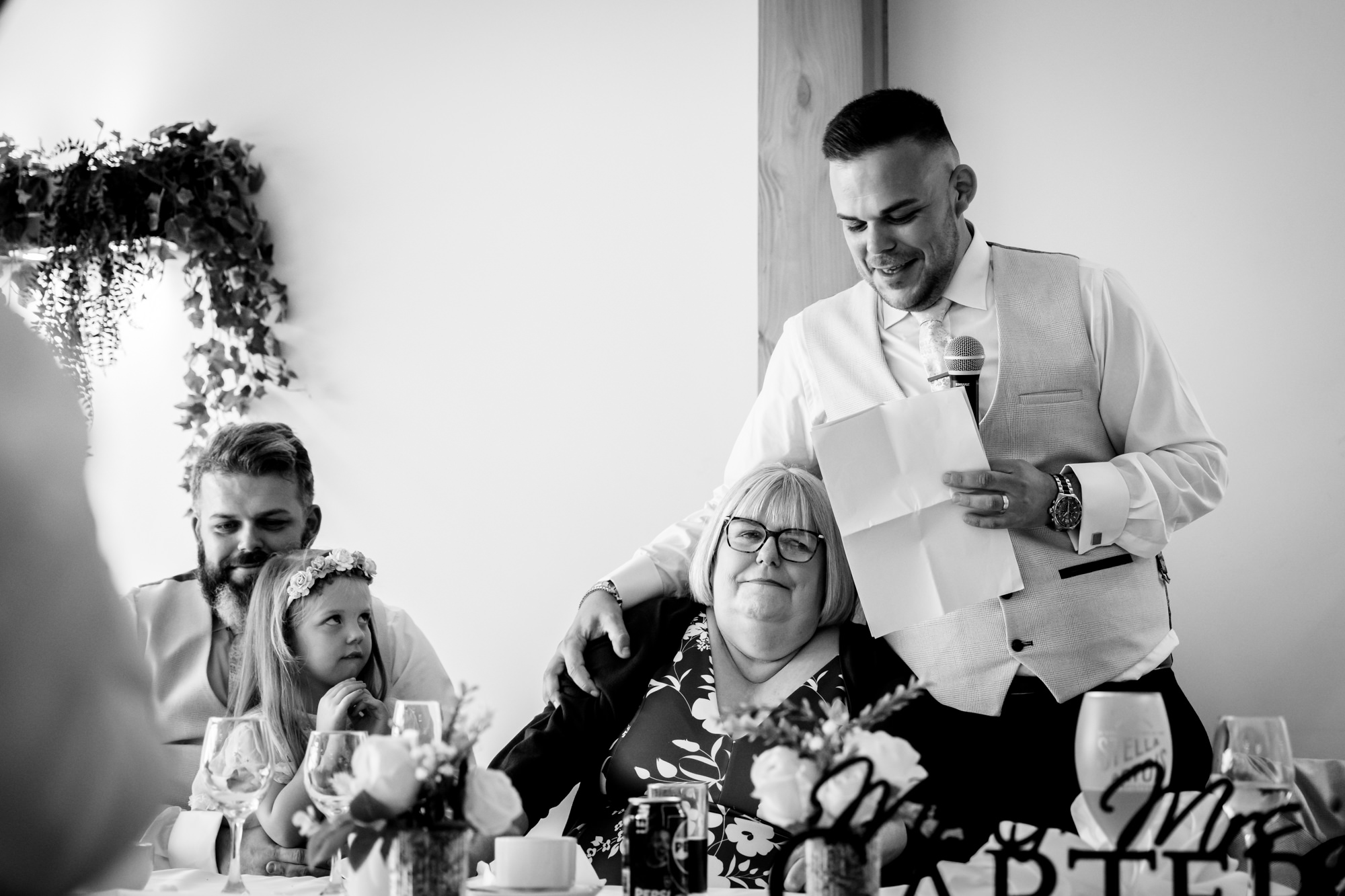The groom standing and giving his speech while holding a microphone, smiling down at his mom at Hanbury Barns.