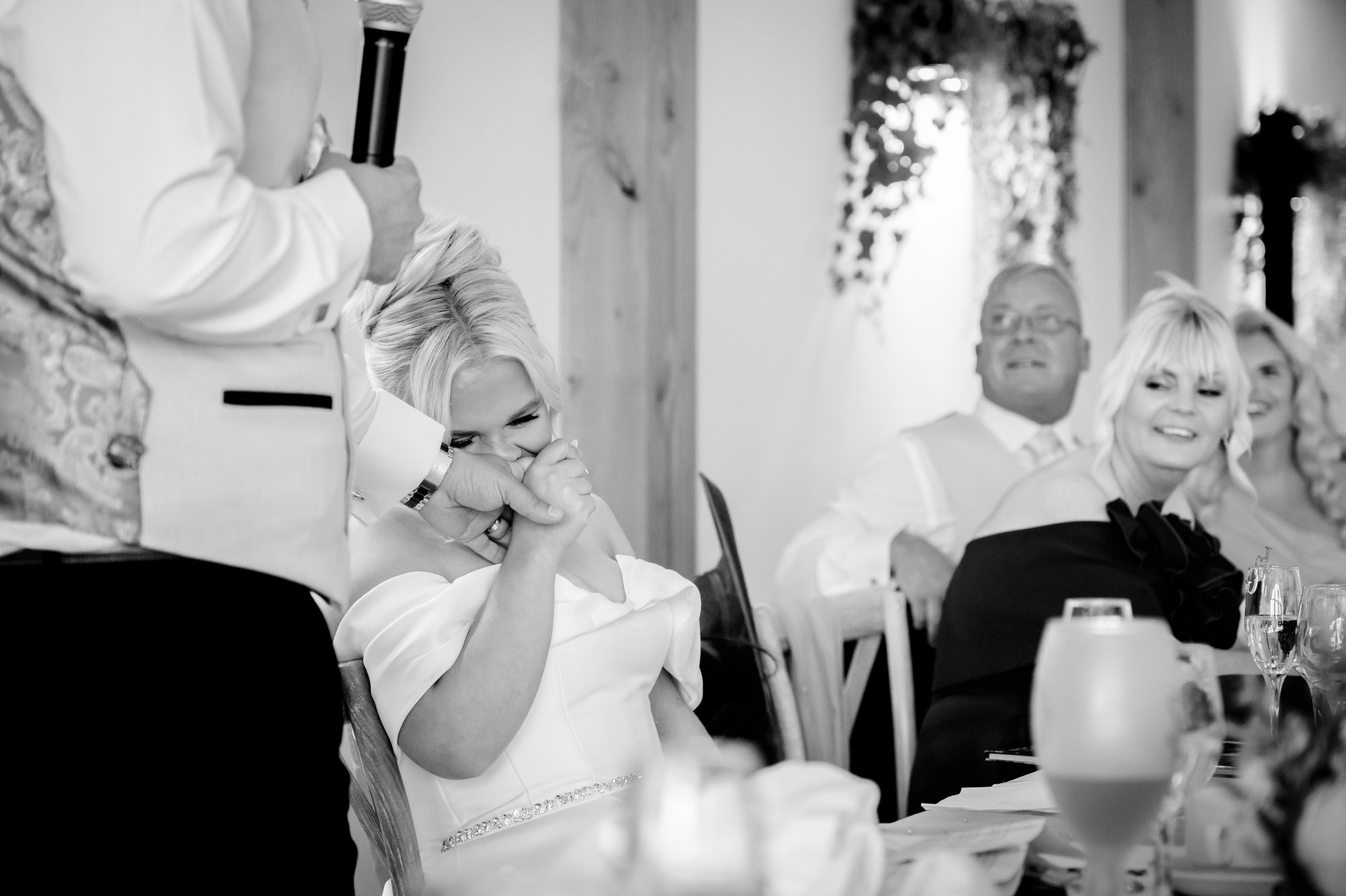 A black and white candid photo of guests at the wedding breakfast table reacting and laughing during a speech at Hanbury Barns.