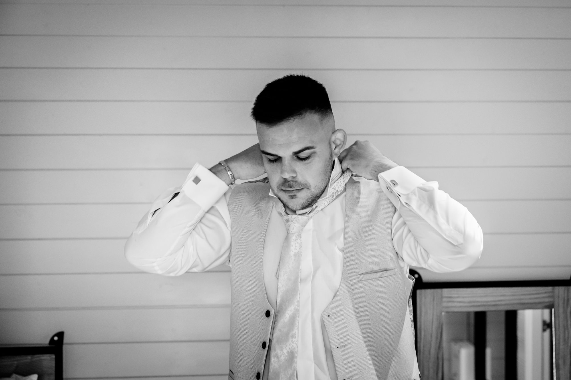 The groom putting on his waistcoat and tying his necktie in black and white inside the curved preparation pod at Hanbury Barns.