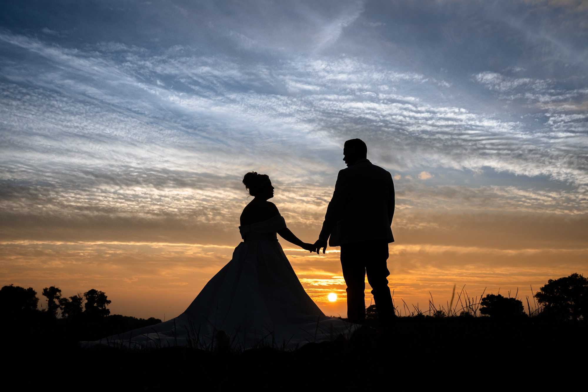 A gorgeous wedding portrait at Hanbury Barns showing the couple embracing in a dark, atmospheric shot with a sunset behind