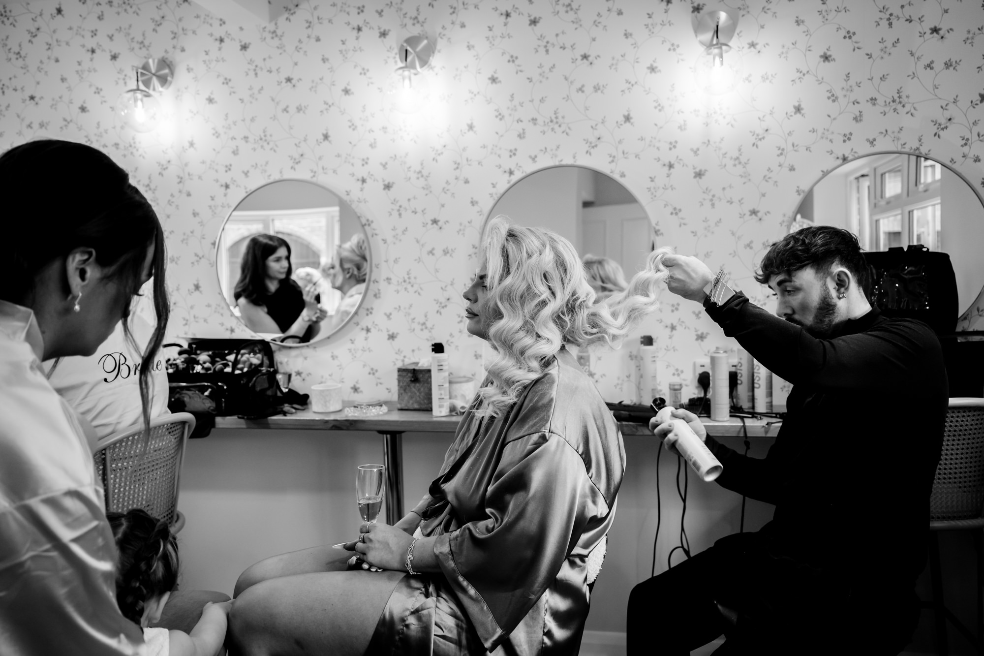 The bridal party and stylists focused on the bridesmaid’s hair and makeup in a black and white shot of the busy dressing room at Hanbury Barns.