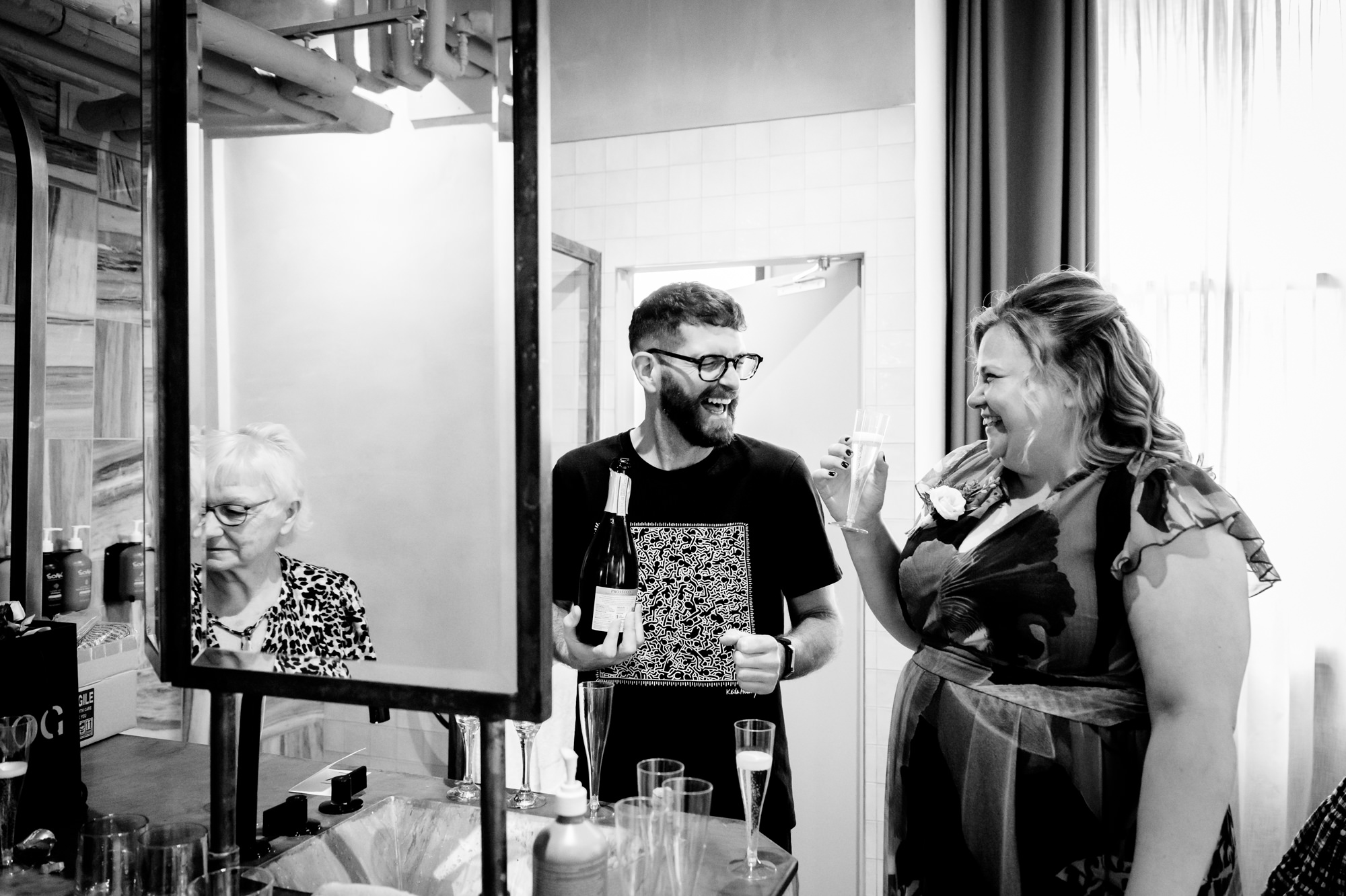 The dark-haired groom laughs happily, holding a bottle of champagne with a guest, with another guest visible in a mirror reflection, at The Alan hotel wedding prep.