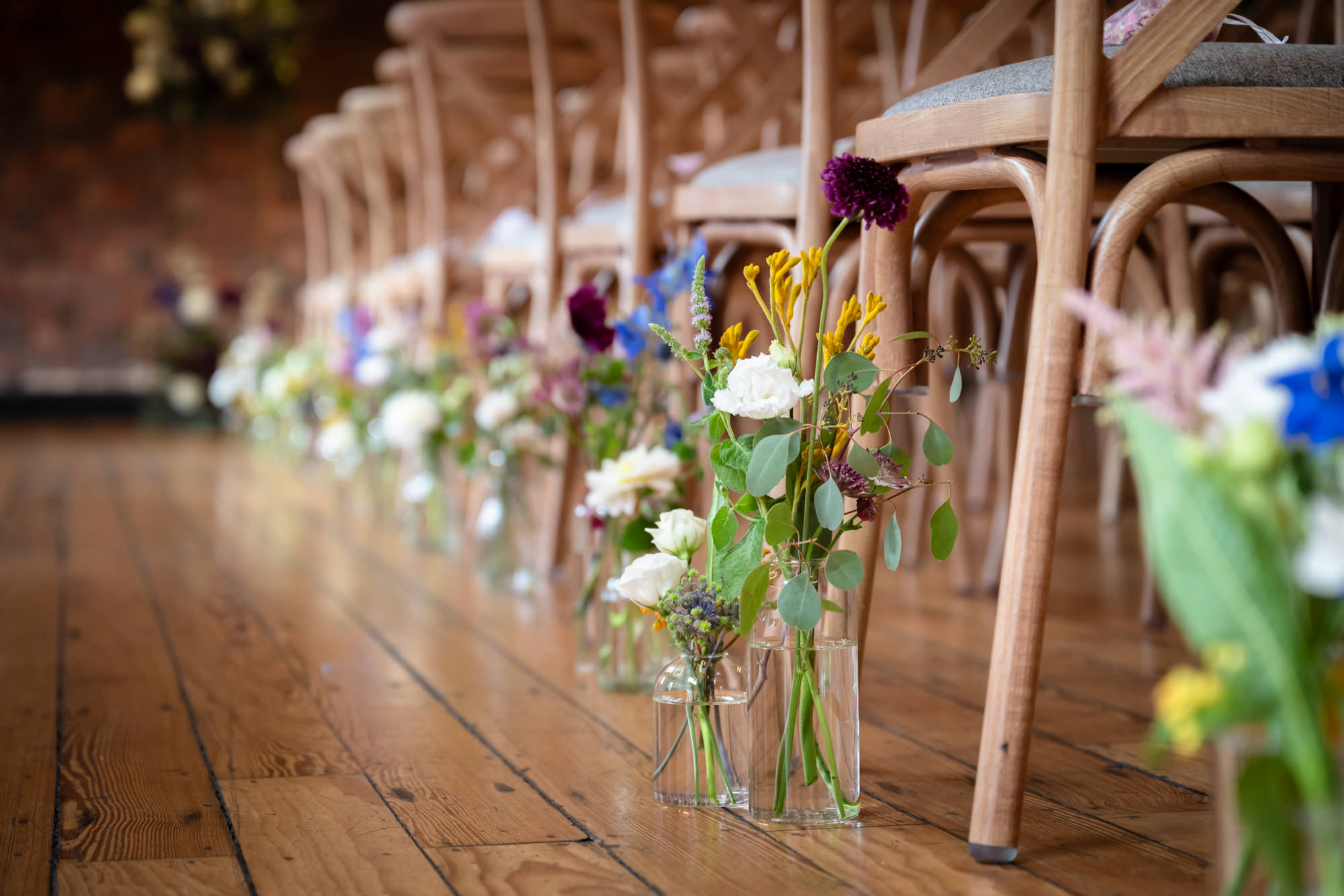 The Brindley room at Castlefield Rooms is ready for the wedding ceremony, featuring rustic wooden cross-back chairs, exposed brick, and floral aisle decorations in jars.