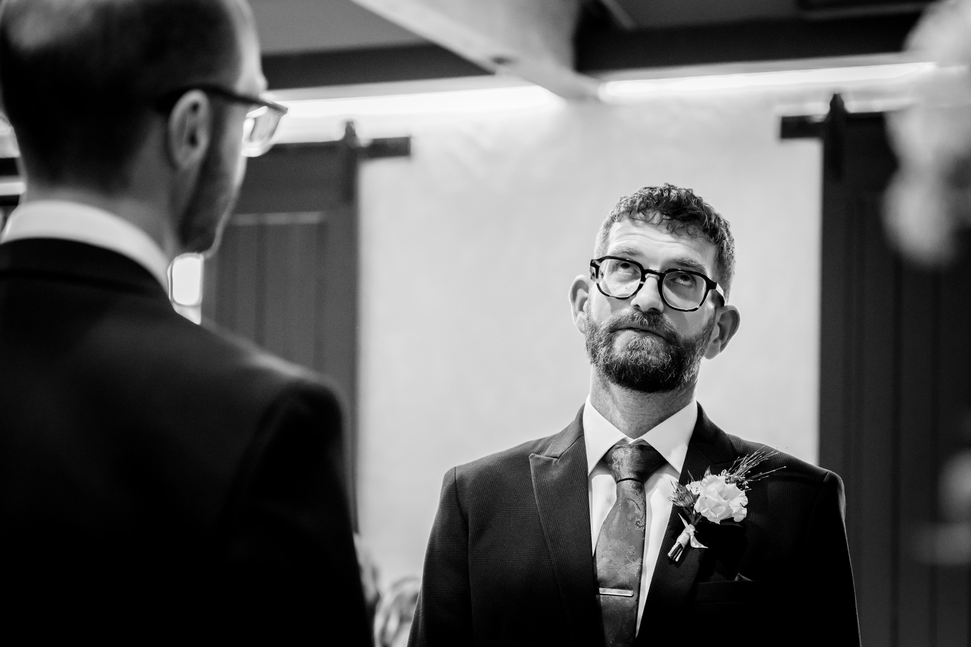The dark-haired groom and bald groom, both in dark suits, wait casually as guests arrive for the wedding ceremony at Castlefield Rooms.