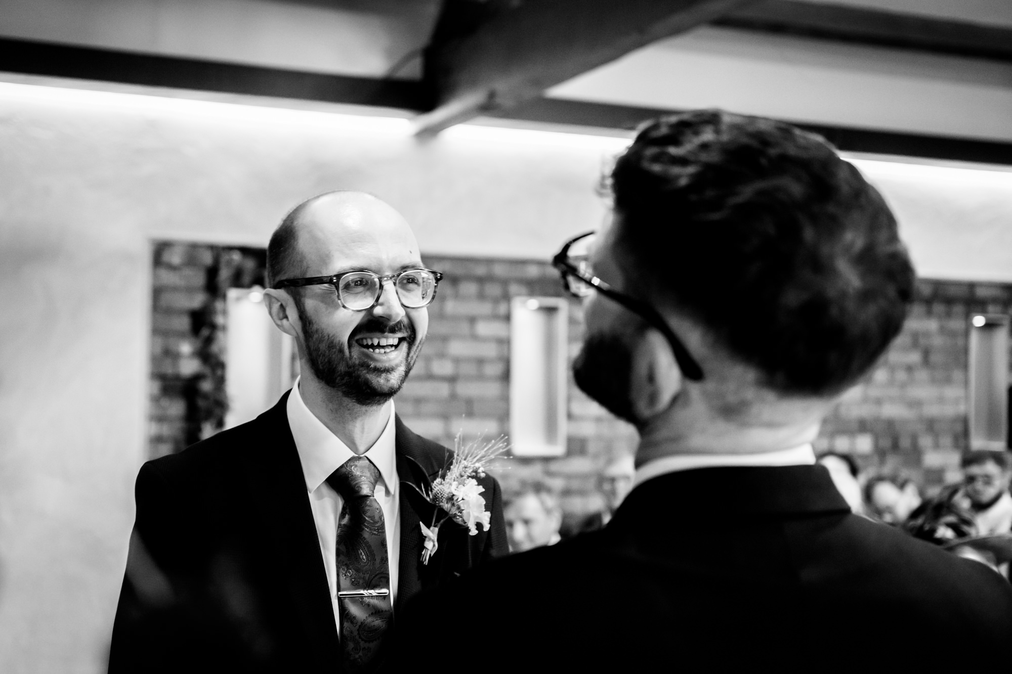 Black and white photo of the dark-haired groom in his blue suit and purple tie, standing and smiling as he awaits the start of the ceremony in the Brindley room at Castlefield Rooms.