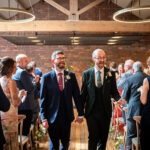 The two smiling grooms walk hand-in-hand down the flower-lined aisle after their ceremony in the Brindley room at Castlefield Rooms.