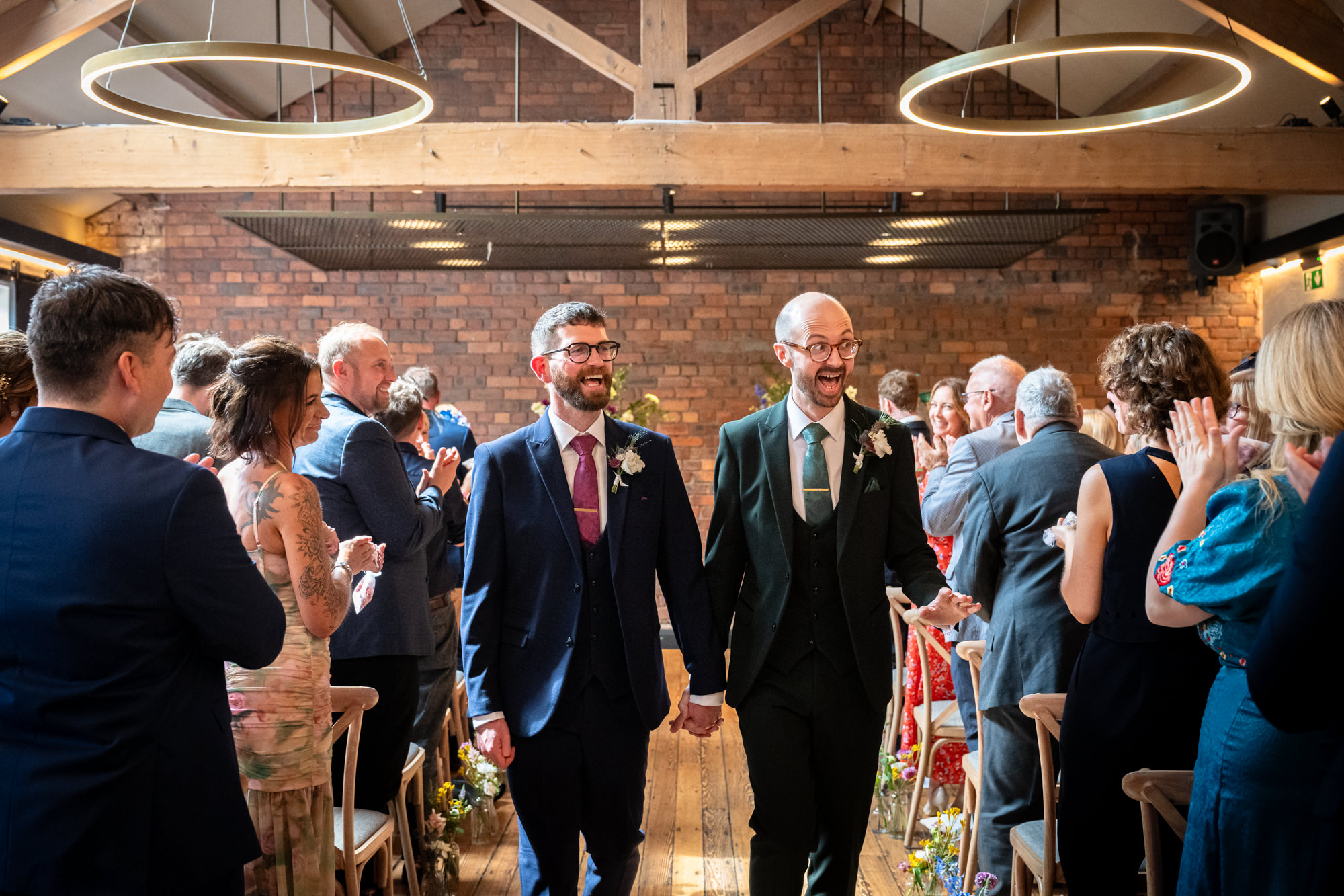 The two smiling grooms walk hand-in-hand down the flower-lined aisle after their ceremony in the Brindley room at Castlefield Rooms.