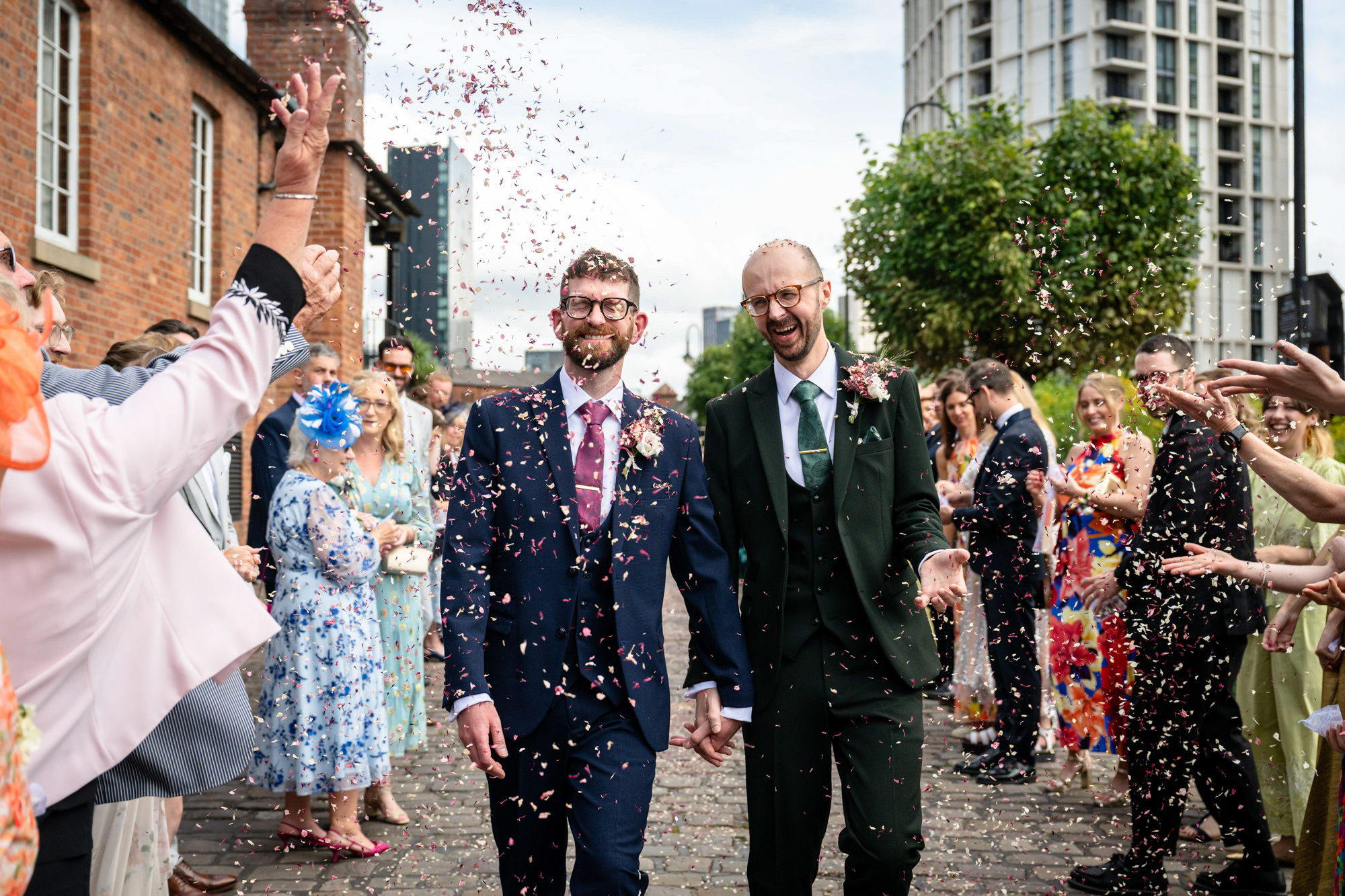 Wedding at Castlefield Rooms. The grooms are showered with confetti by their delighted guests as they exit the venue, holding hands and laughing.