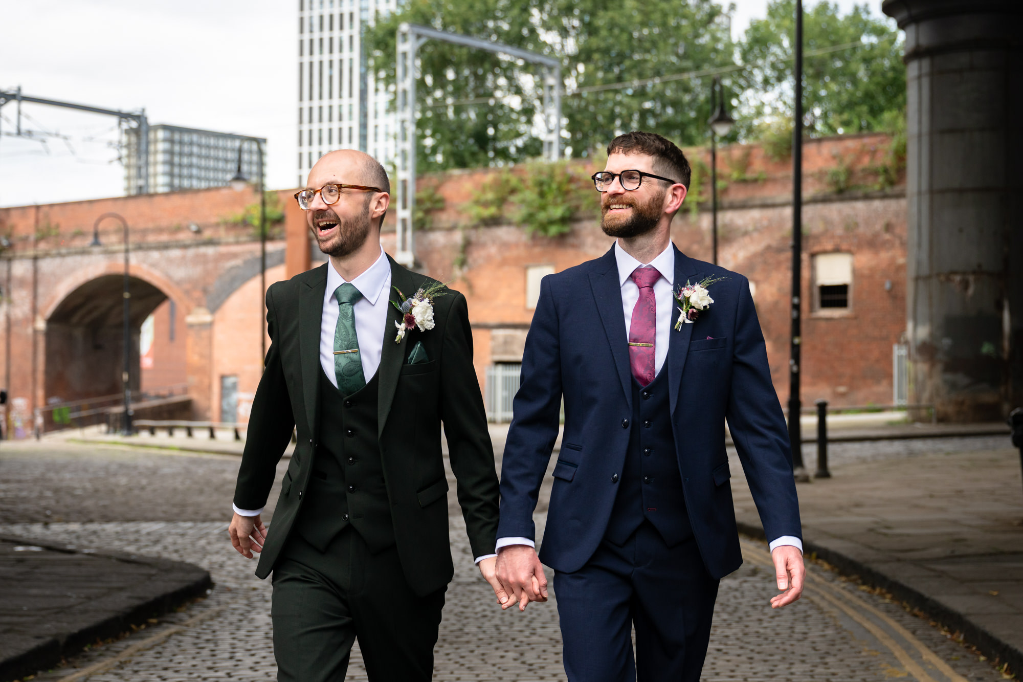 A dramatic, wide black and white portrait of the two grooms standing beneath the towering arches of the Castlefield viaducts.