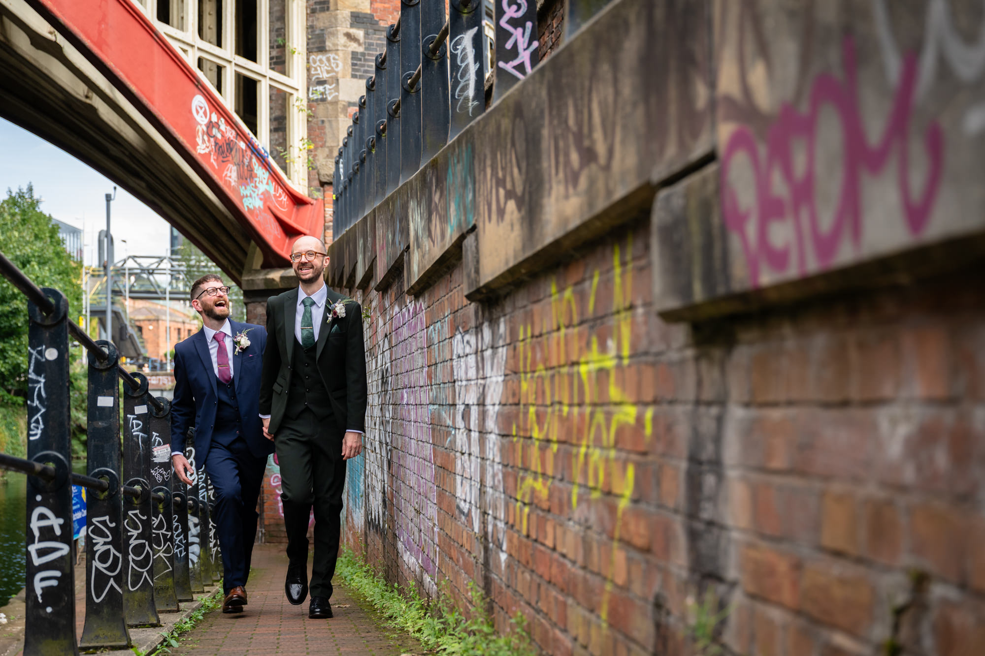 The two grooms walk hand-in-hand and smile at each other during their portrait session in the industrial urban setting near Castlefield Rooms.