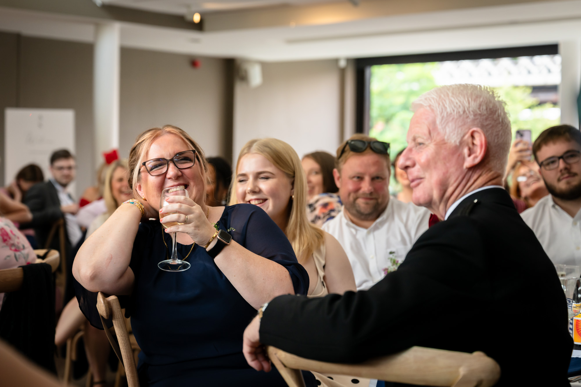 The groom in the green suit smiles from the top table, surrounded by beautiful flowers, listening intently to the speeches at the Castlefield Rooms wedding reception.