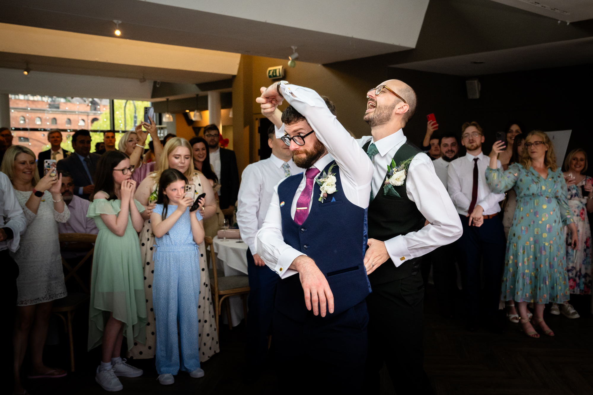 A slightly wider shot of the grooms standing to give their joint speech to all the seated wedding guests at the Castlefield Rooms reception.