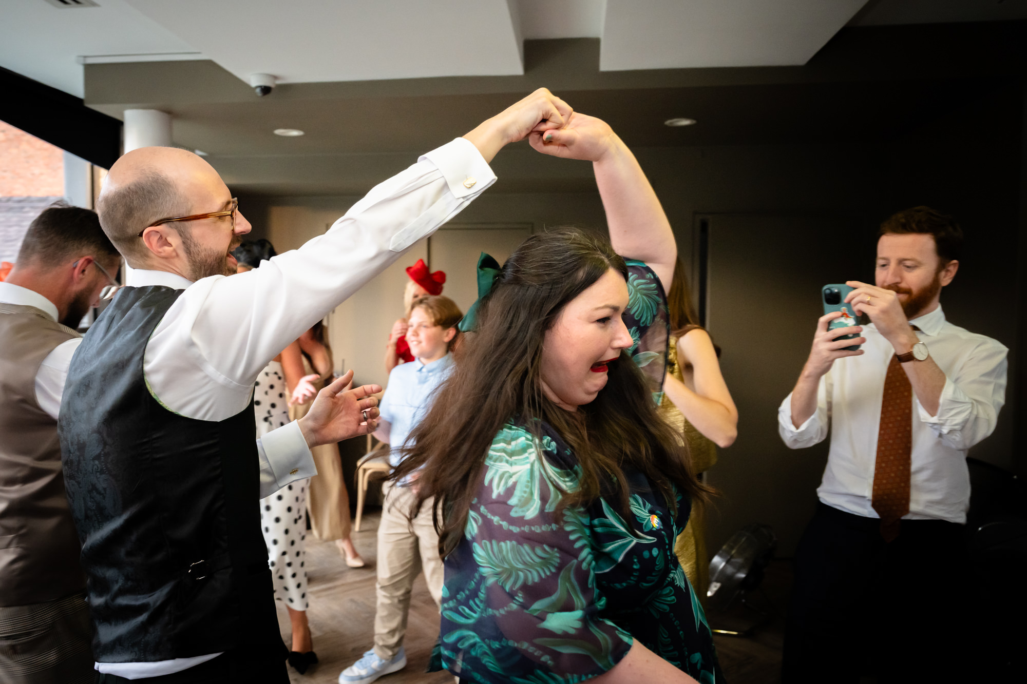 The groom in the blue suit raises his hand in the air, singing and dancing enthusiastically surrounded by friends at the evening reception at Castlefield Rooms.