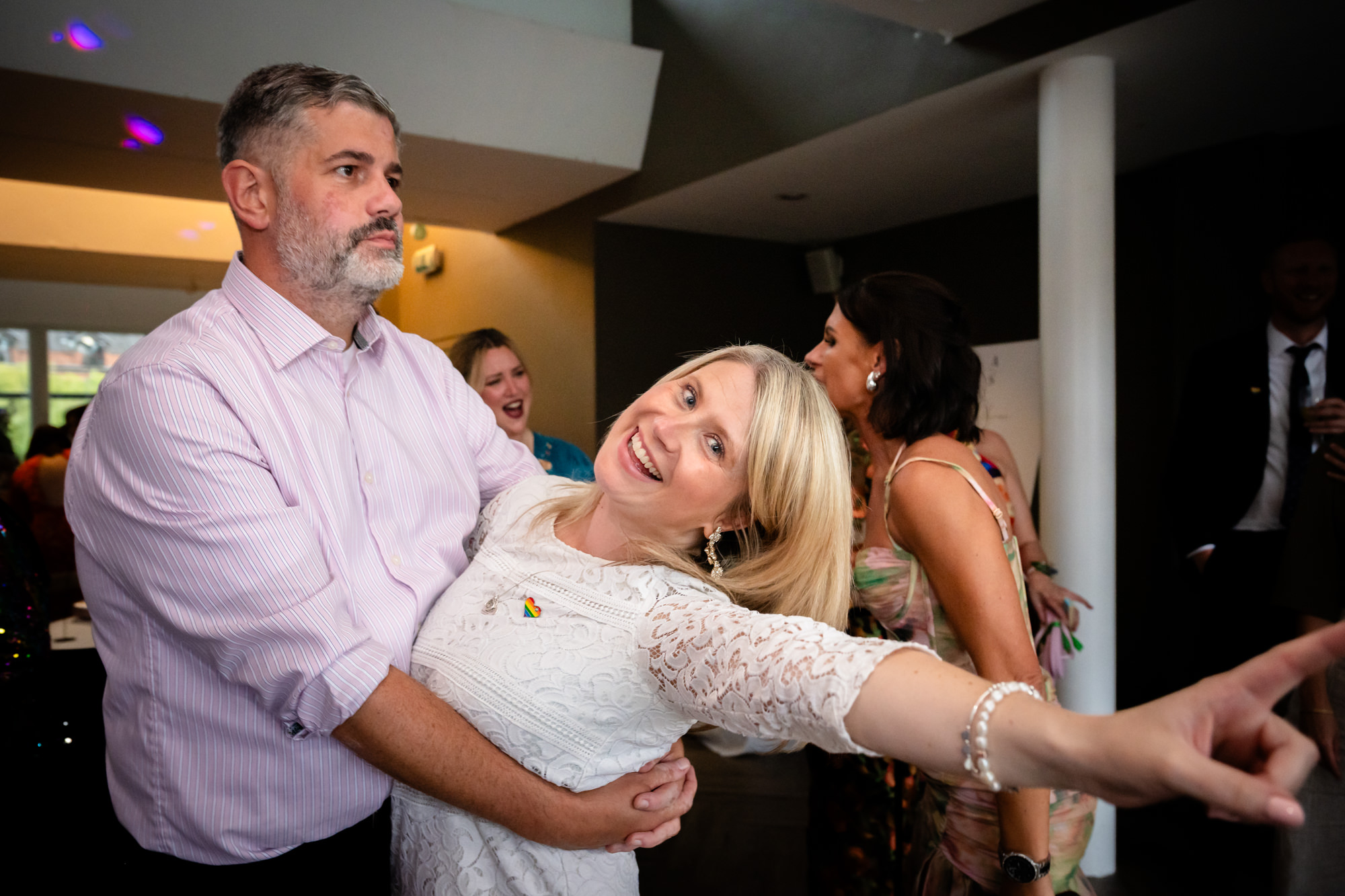 The groom in the blue suit is surrounded by dancing guests, raising his hands in celebration during the evening party at Castlefield Rooms.