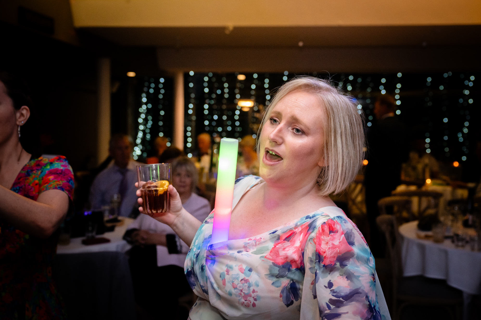 The groom in the green suit throws his arms up with glow sticks, celebrating an upbeat moment on the dance floor at the Castlefield Rooms evening party.