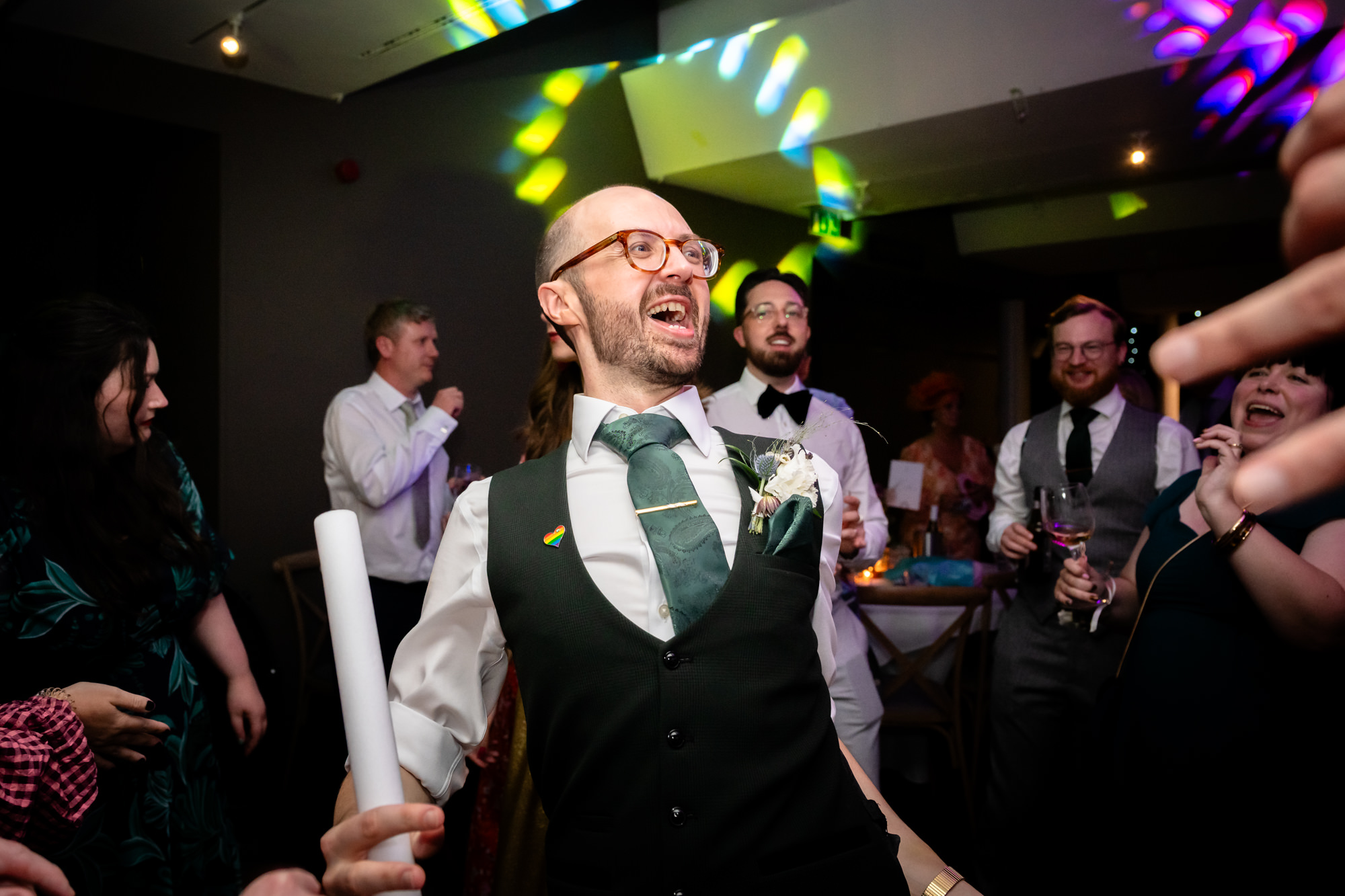 The groom in the green suit, full of energy and smiling, dances with a glow stick and is surrounded by guests at the lively evening party at Castlefield Rooms.