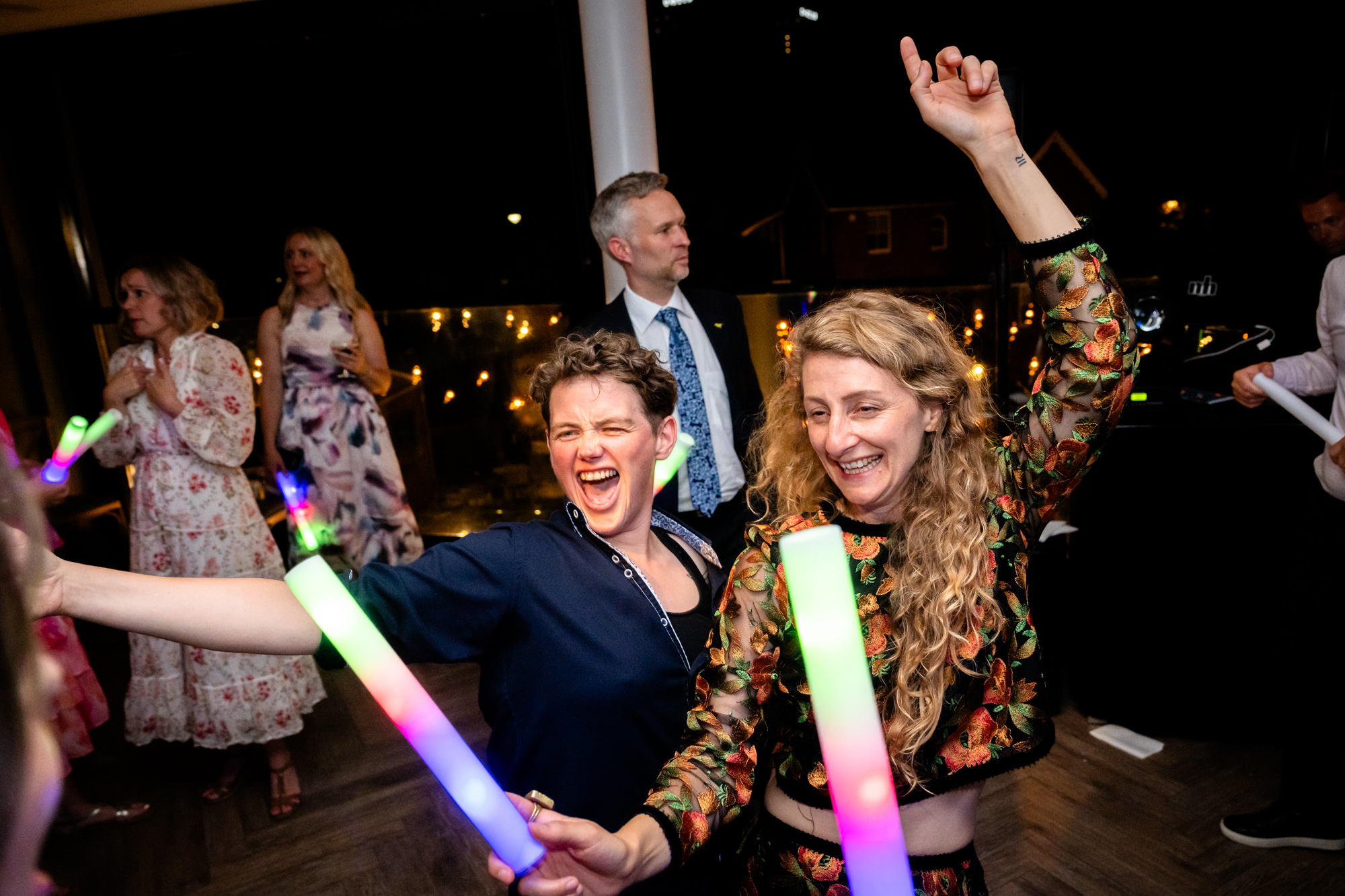 Two female guests, laughing and holding glow sticks, pose for a photo on the dance floor during the upbeat party at Castlefield Rooms.