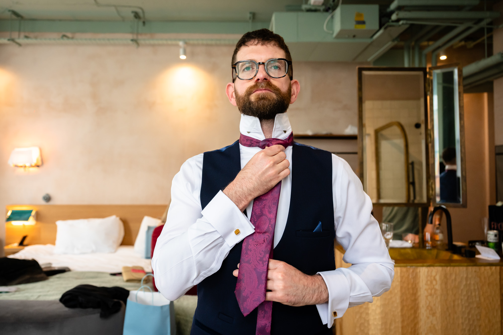 The bald groom, smiling and wearing glasses, adjusts the cuff of his white shirt near a bright window at The Alan hotel before the Castlefield Rooms wedding.