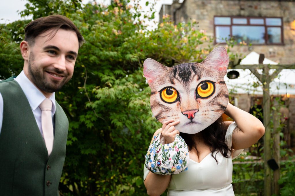The bride playfully holds a large, funny cat mask over her face as the groom smiles next to her at their Roman Lakes wedding.
