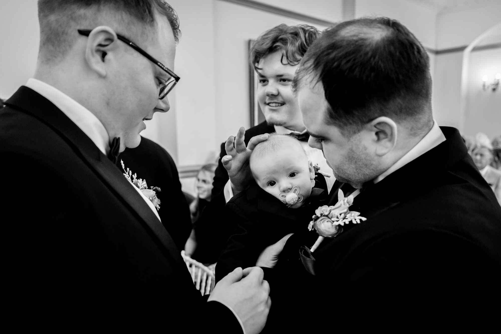 The groom waiting nervously at the front of the aisle at Nunsmere Hall holding a baby in a black suit.