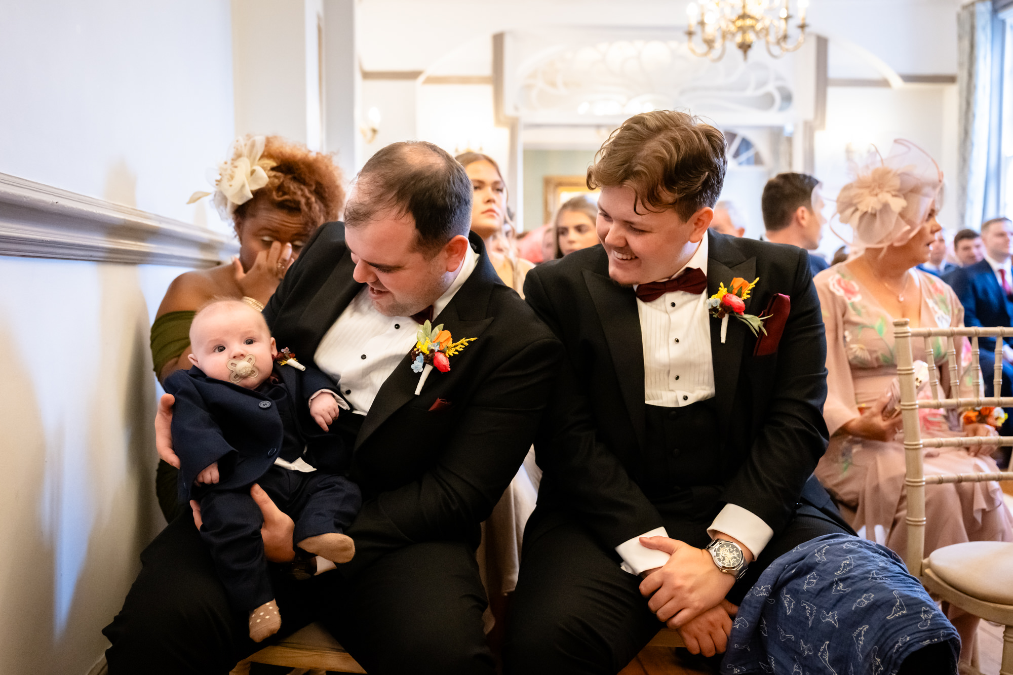 A guest holding a baby in a suit while seated for the wedding ceremony at Nunsmere Hall.