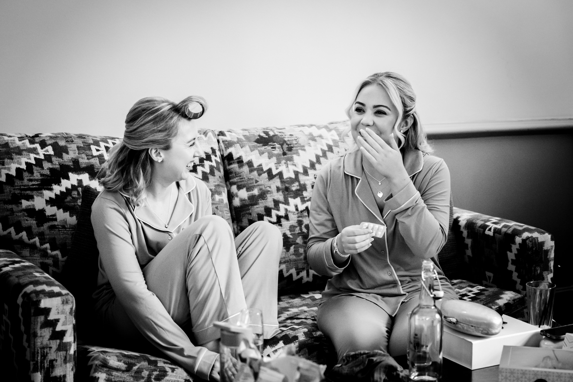 Two bridesmaids in matching pyjamas sitting on a sofa laughing together during morning preparations at Nunsmere Hall.