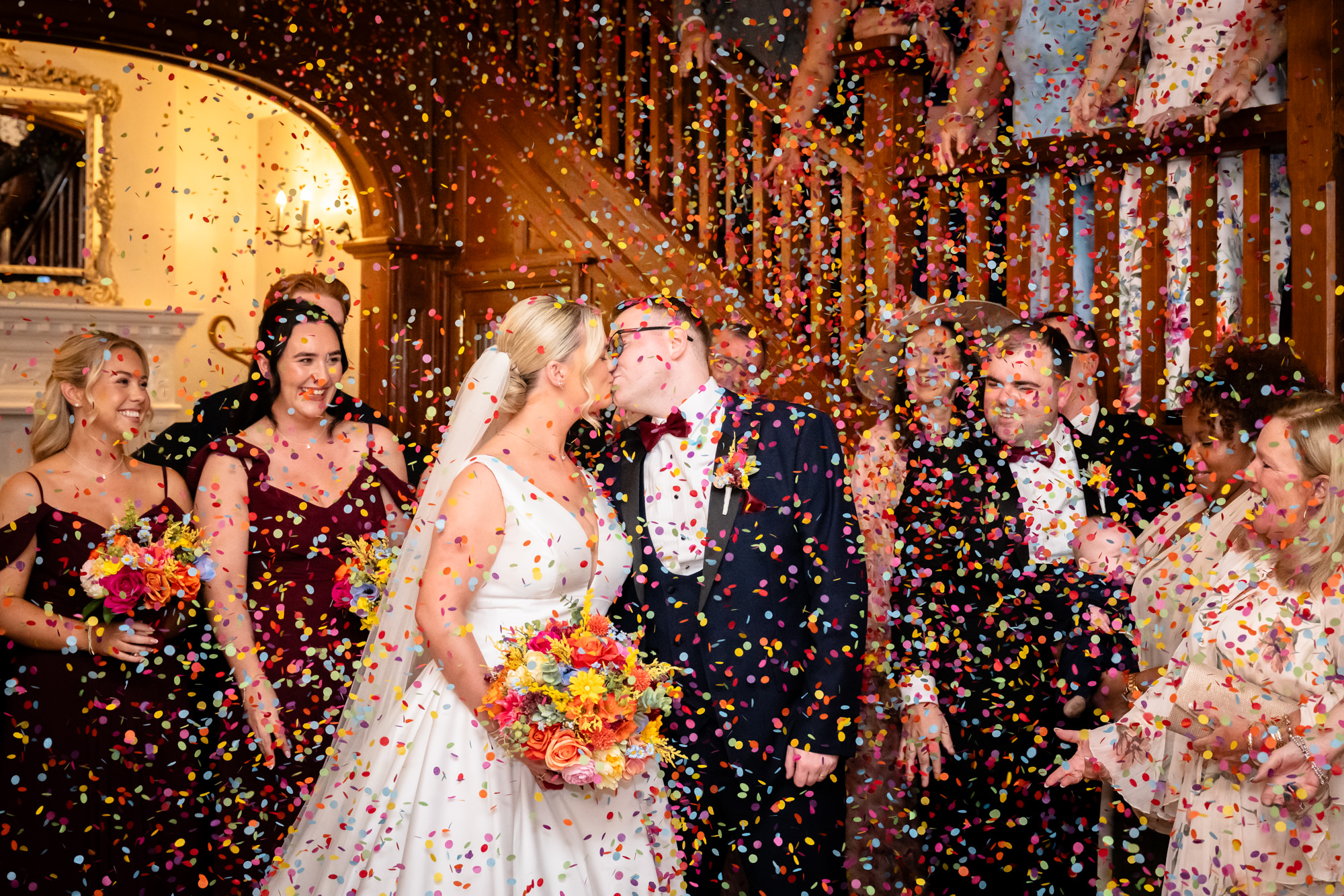 The newlyweds kissing on the stairs while guests shower them with confetti during a wedding at Nunsmere Hall.