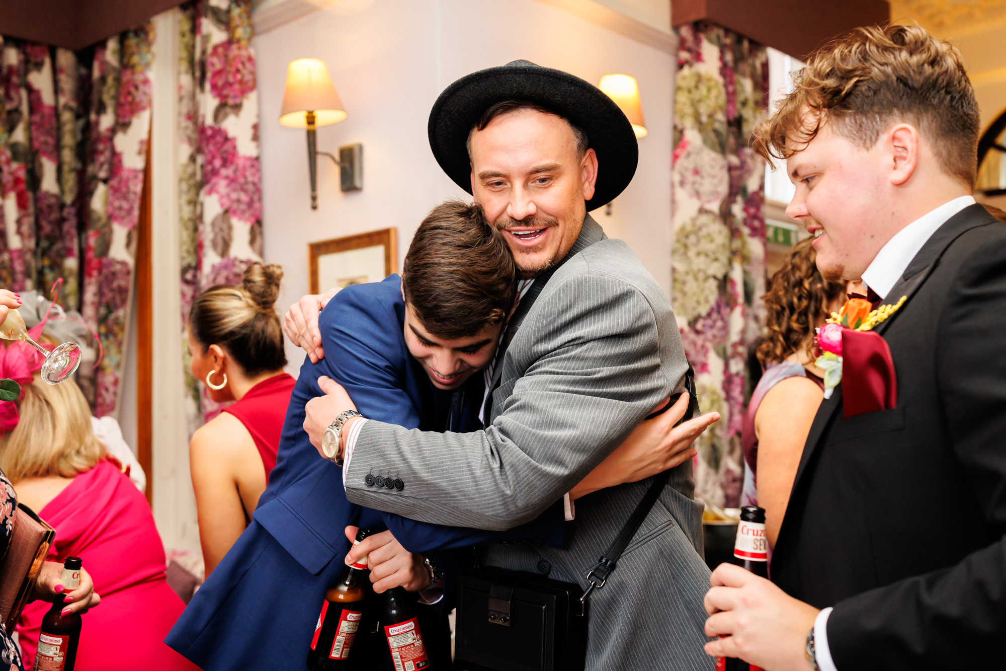 The groom hugging a guest wearing a hat and blue suit at Nunsmere Hall.