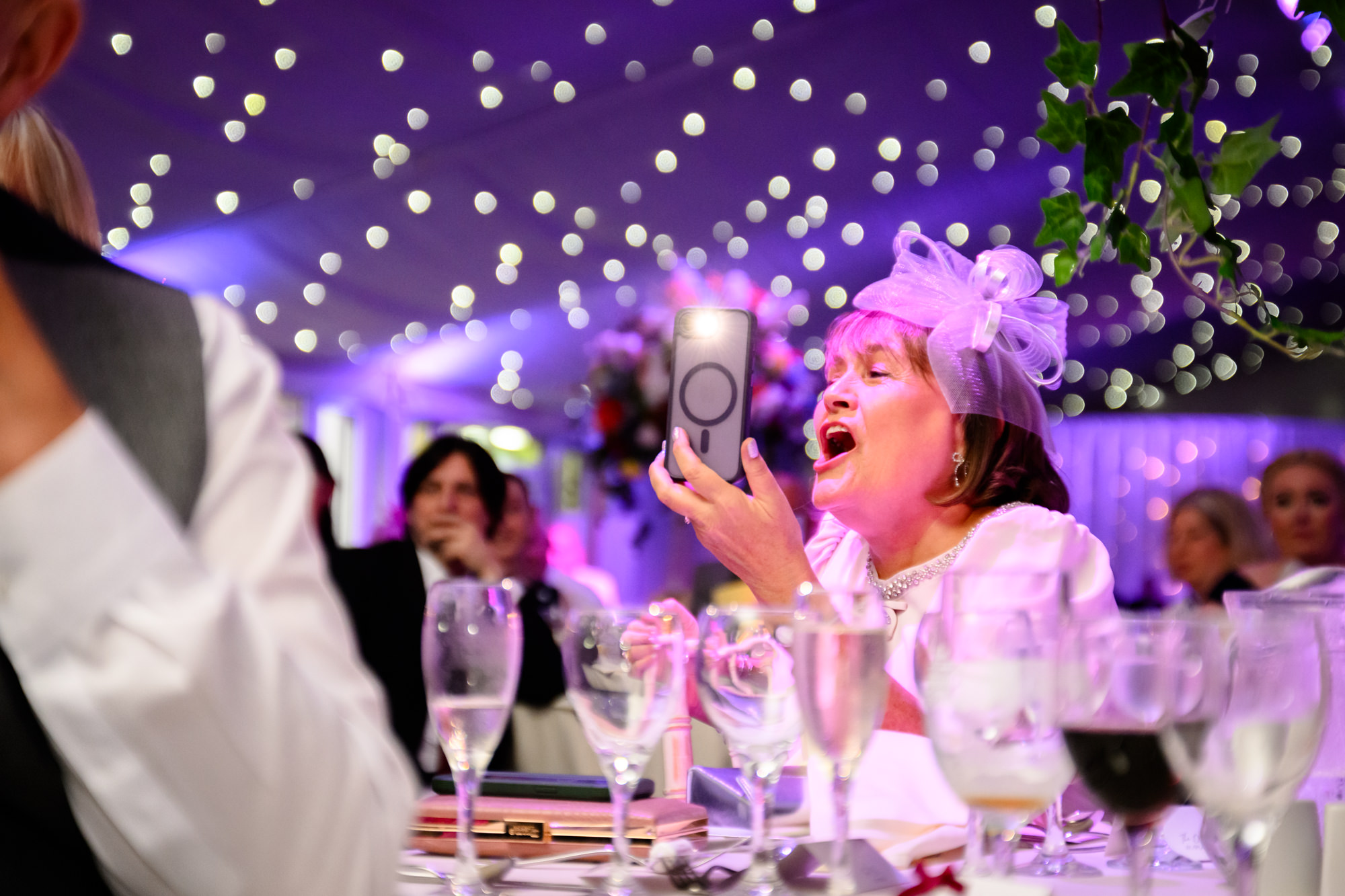 A guest in a pink hat laughing while filming the speeches on her phone at Nunsmere Hall.