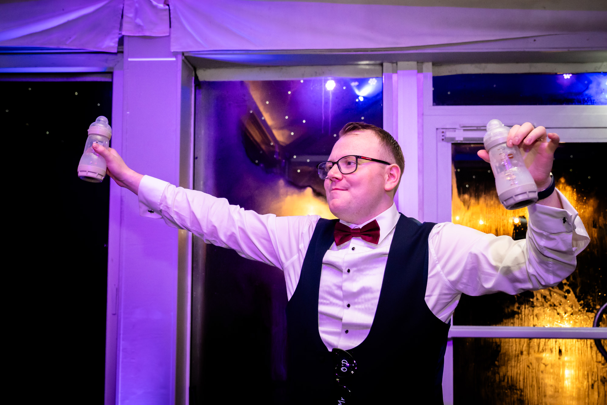 The groom holding two baby bottles and smiling widely during the party at Nunsmere Hall.