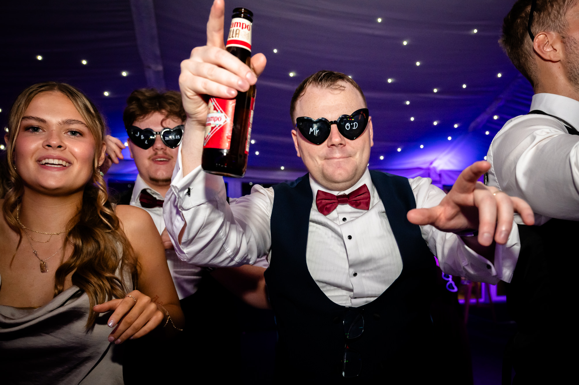 The groom wearing sunglasses holding a bottle of beer and dancing at a wedding at Nunsmere Hall.