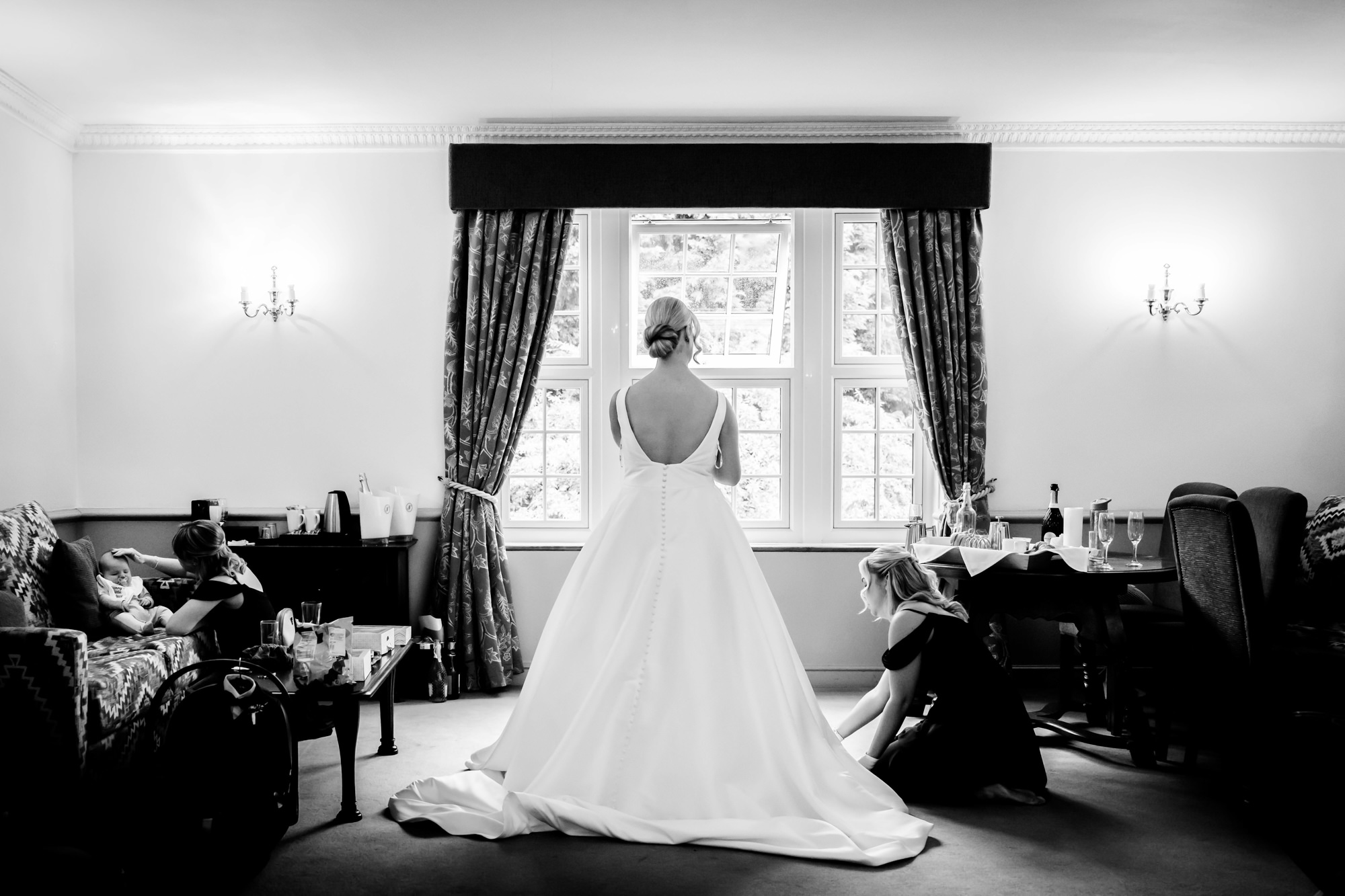 The bride stands by a large window looking out at the grounds before the wedding at Nunsmere Hall while bridesmaids adjust her train.