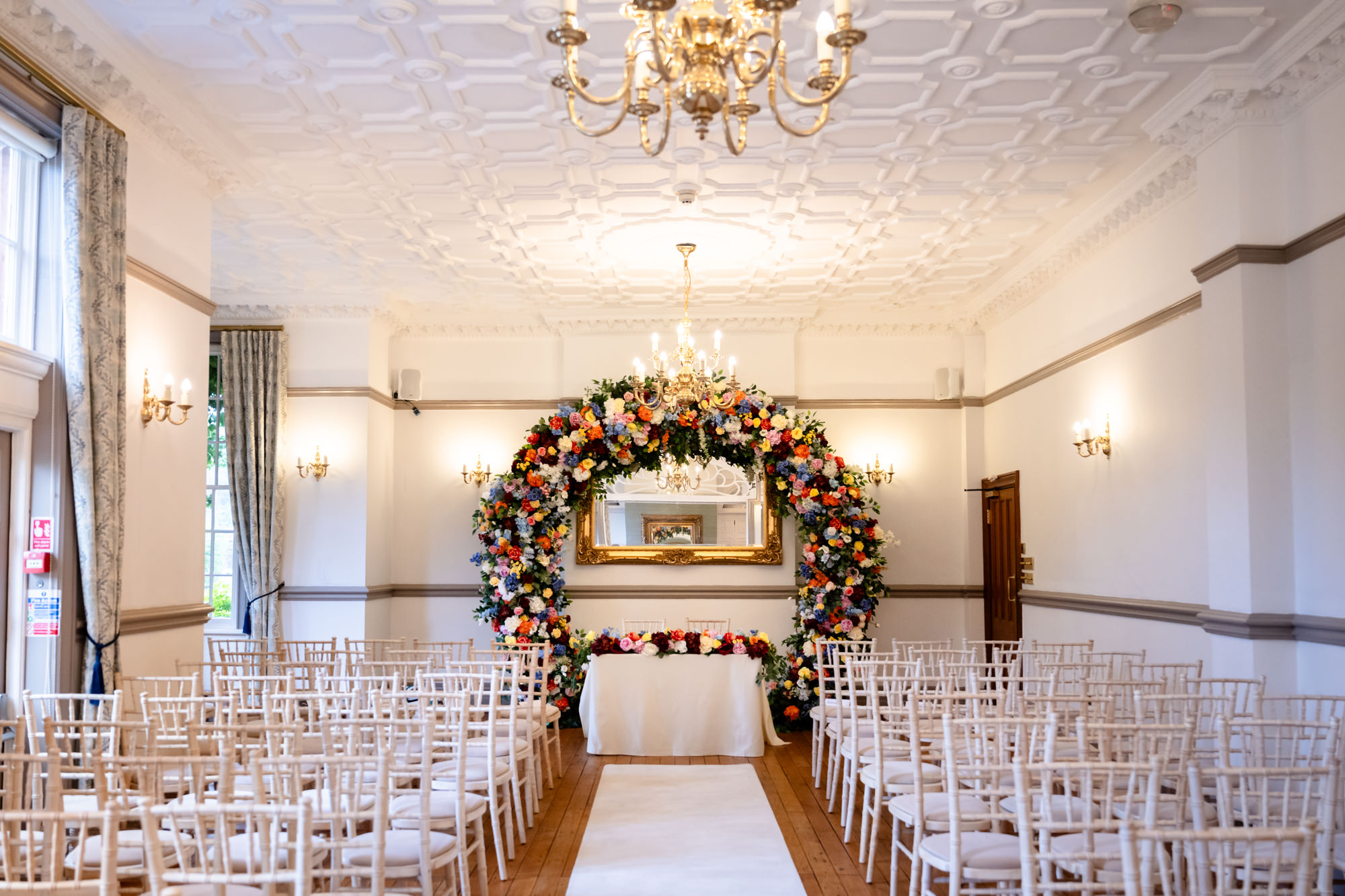 The Crystal Suite ready for a wedding at Nunsmere Hall featuring a white aisle runner and a large colourful floral arch.