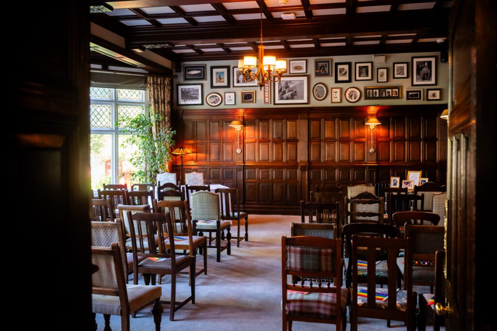 The ceremony room at The Oakfield Chester Zoo set up for a wedding, with rows of mismatched wooden chairs, dark wood panelling and a gallery wall of framed photos