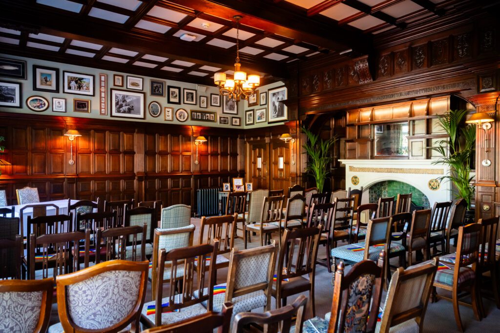 The Oakfield ceremony room at Chester Zoo set for a wedding, showing the original fireplace with green tiles, wood panelling, carved ceiling and Pride ribbon chair decorations