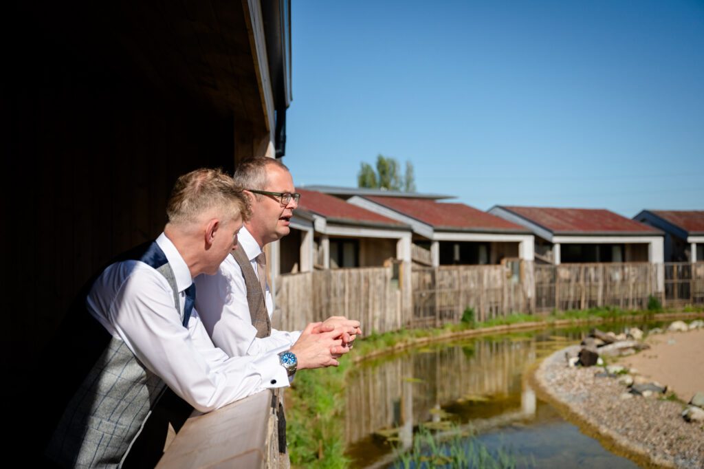 Two grooms in waistcoats leaning on a balcony at The Reserve Chester Zoo, looking at the giraffes their wedding morning