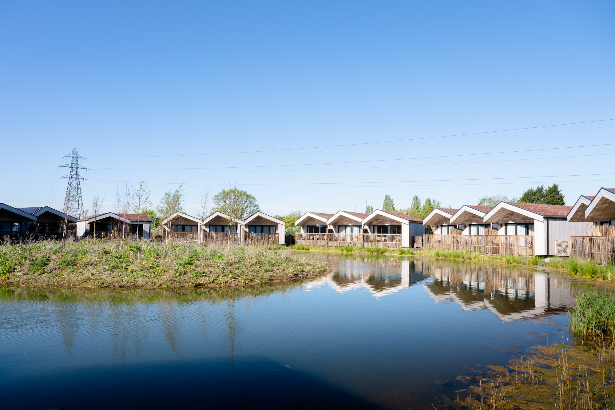 Lakeside lodges at The Reserve Chester Zoo reflected in calm water, with natural vegetation and open sky