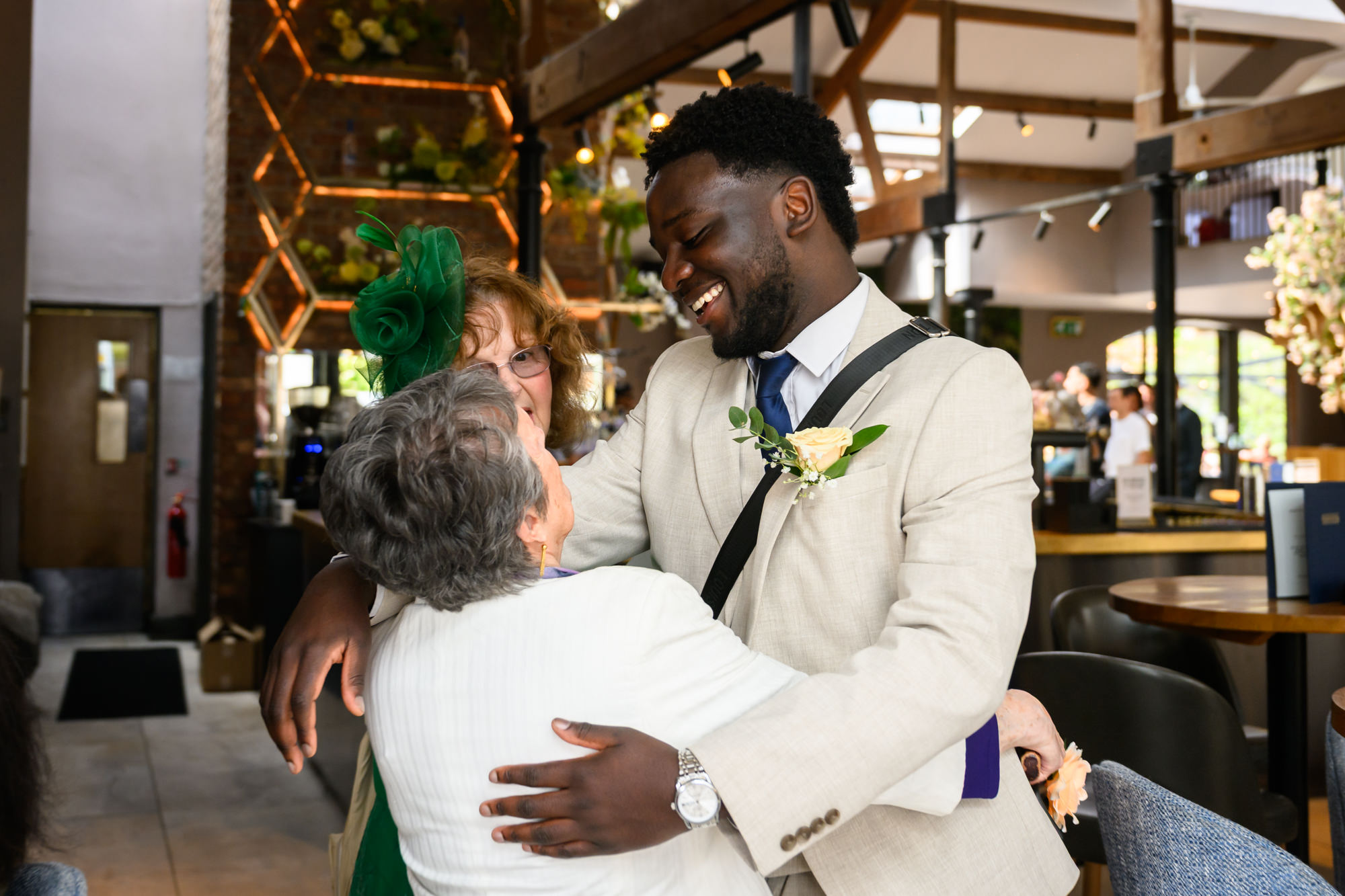 A guest in a cream suit with a yellow rose buttonhole hugging another guest at The Castlefield Rooms before the wedding ceremony.