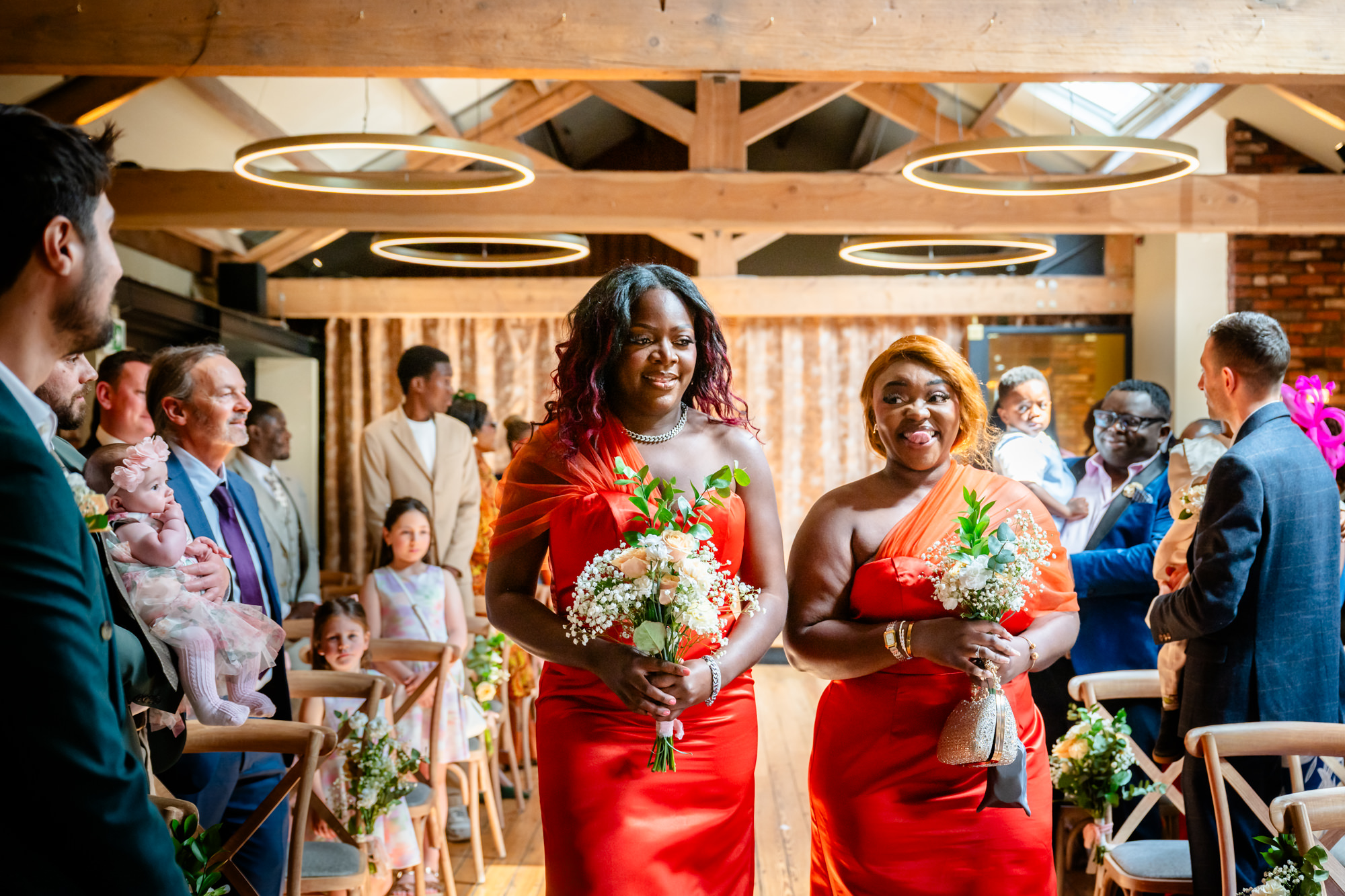 Two bridesmaids in orange dresses walking down the aisle at The Castlefield Rooms during the wedding ceremony, carrying bouquets of peach roses and gypsophila.