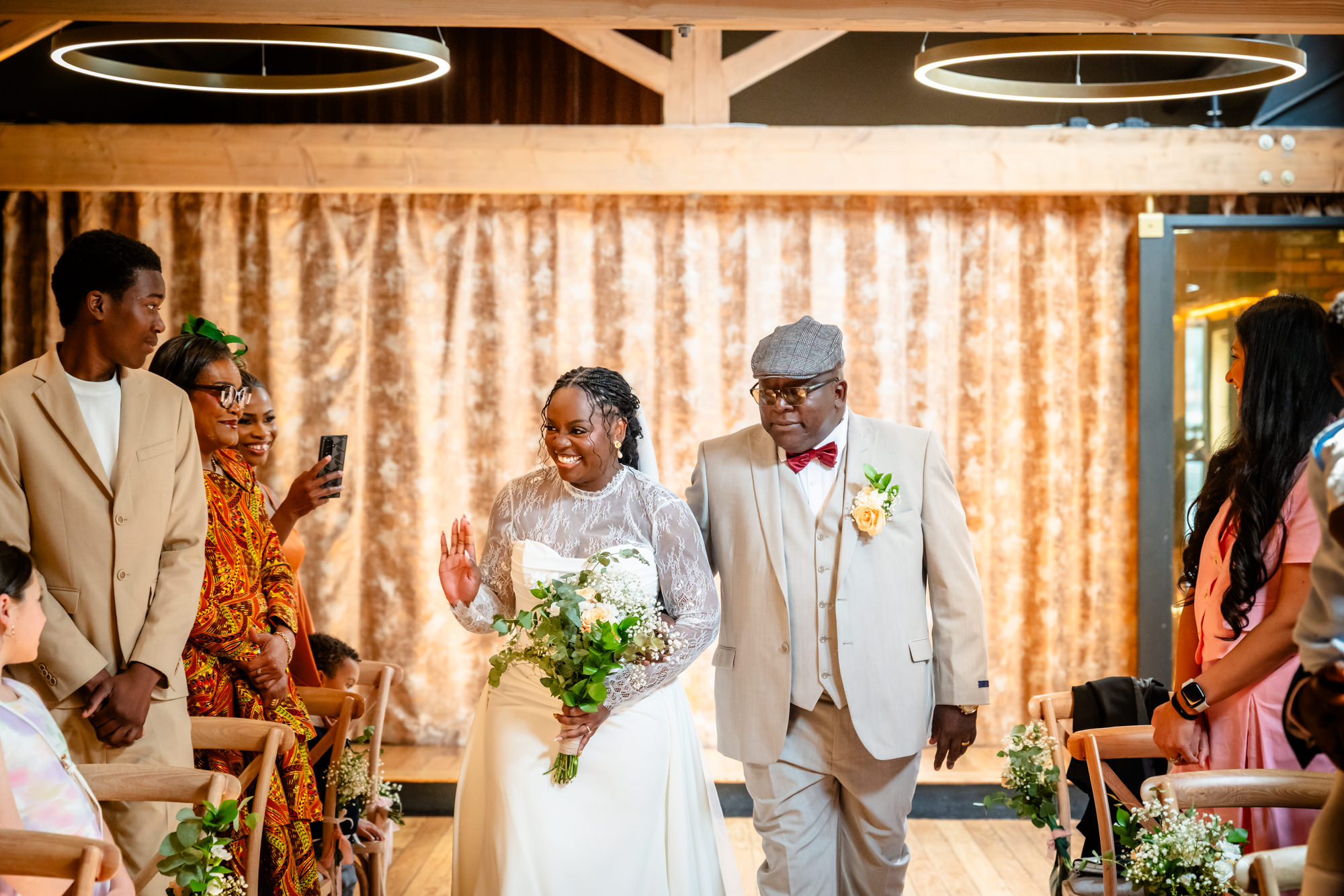The bride walking down the aisle with her father at The Castlefield Rooms, smiling and waving to guests during the wedding ceremony.