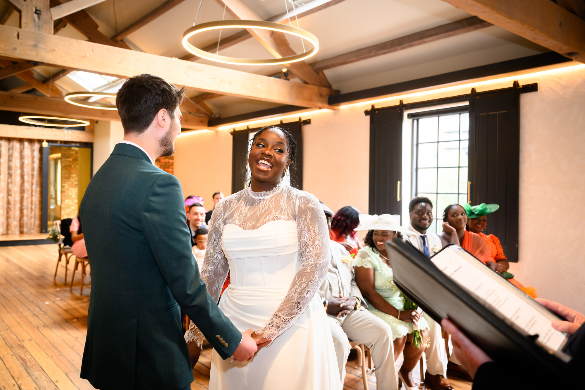 The bride and groom holding hands and smiling during their civil ceremony in the Brindley Room at The Castlefield Rooms, with the registrar visible to the side.
