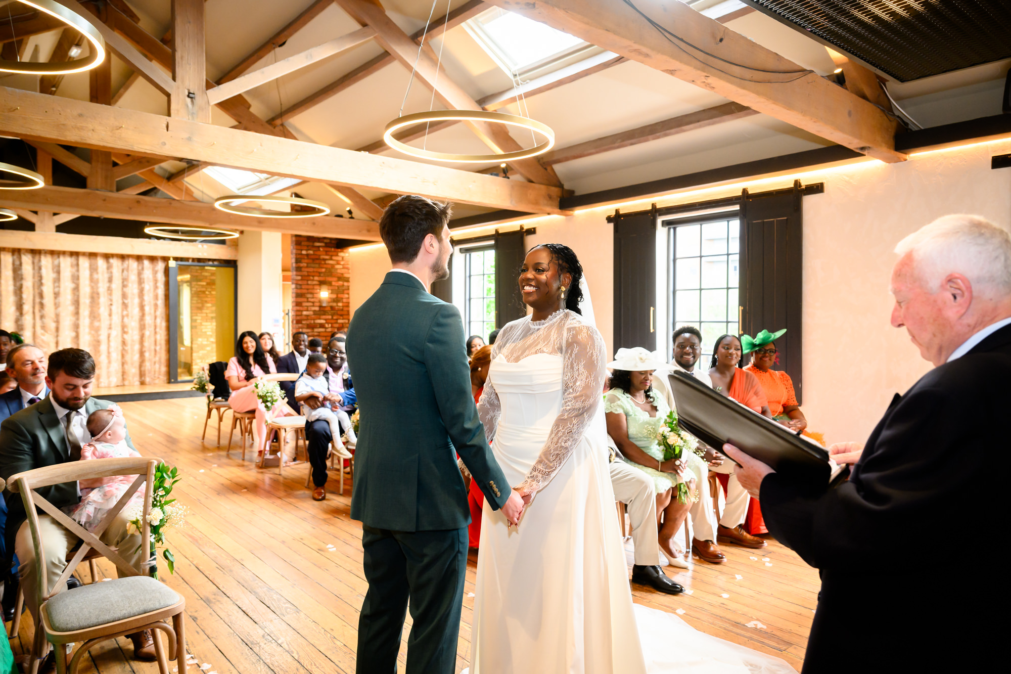 Wide shot of the bride and groom holding hands at the altar in the Brindley Room at The Castlefield Rooms, with the registrar conducting the ceremony and guests watching on.