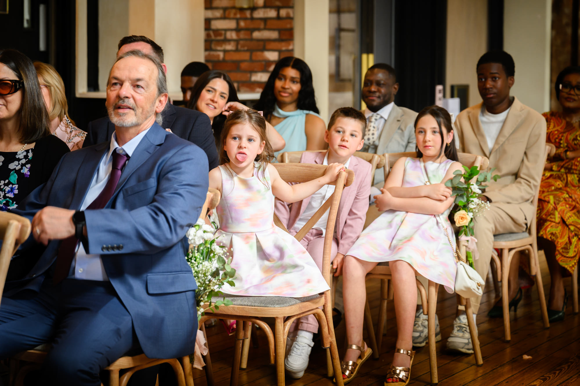 A young flower girl sticking her tongue out during the wedding ceremony in the Brindley Room at The Castlefield Rooms.