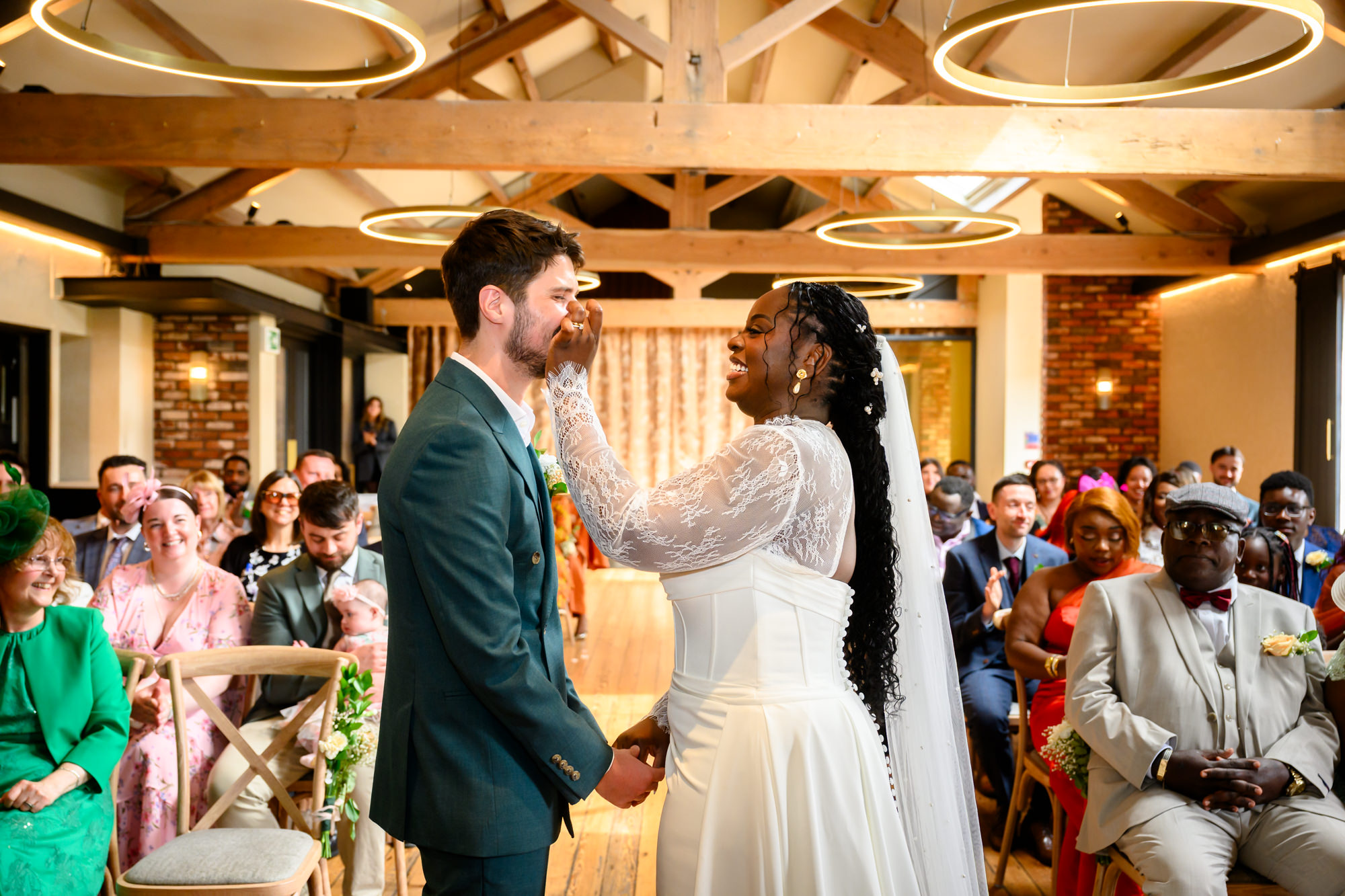The bride wiping a tear from the groom's cheek during their wedding ceremony in the Brindley Room at The Castlefield Rooms, with guests applauding in the background.