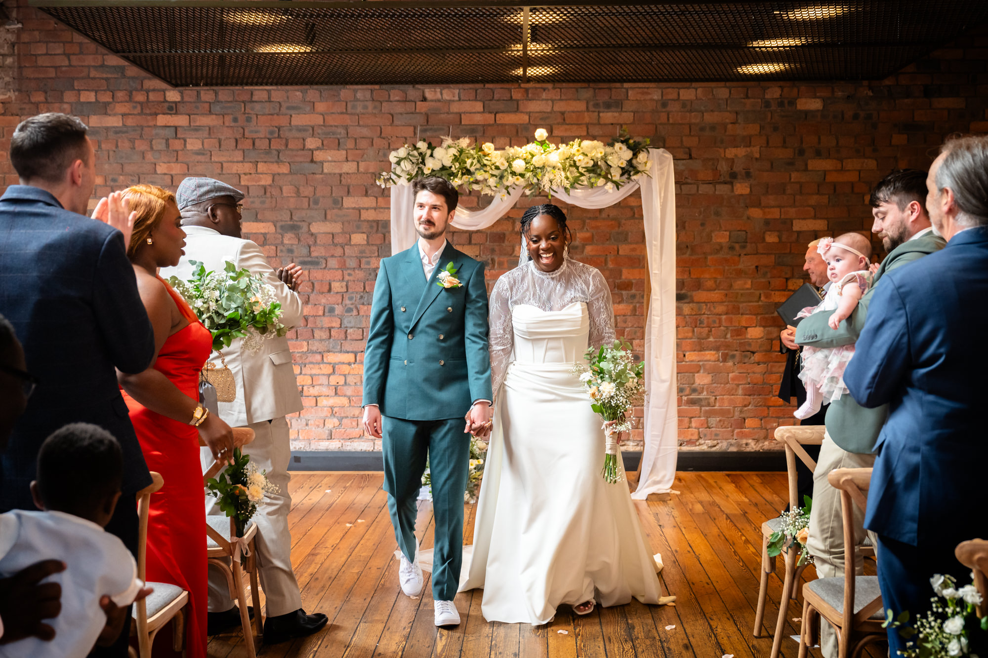The newly married couple walking hand in hand back down the aisle in the Brindley Room at The Castlefield Rooms, with guests cheering on either side.