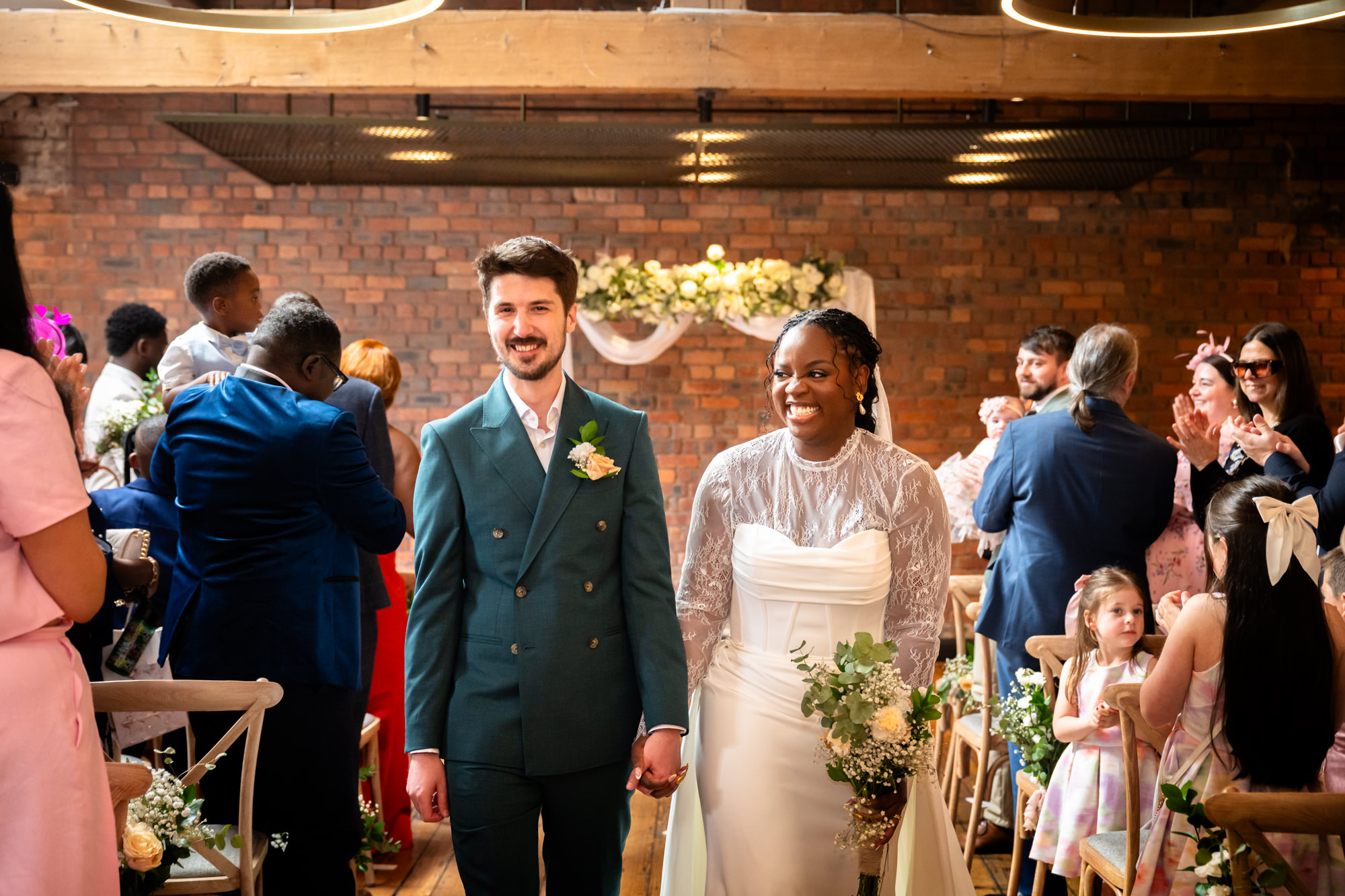 The bride and groom walking back down the aisle at The Castlefield Rooms, both smiling, with guests applauding on either side.